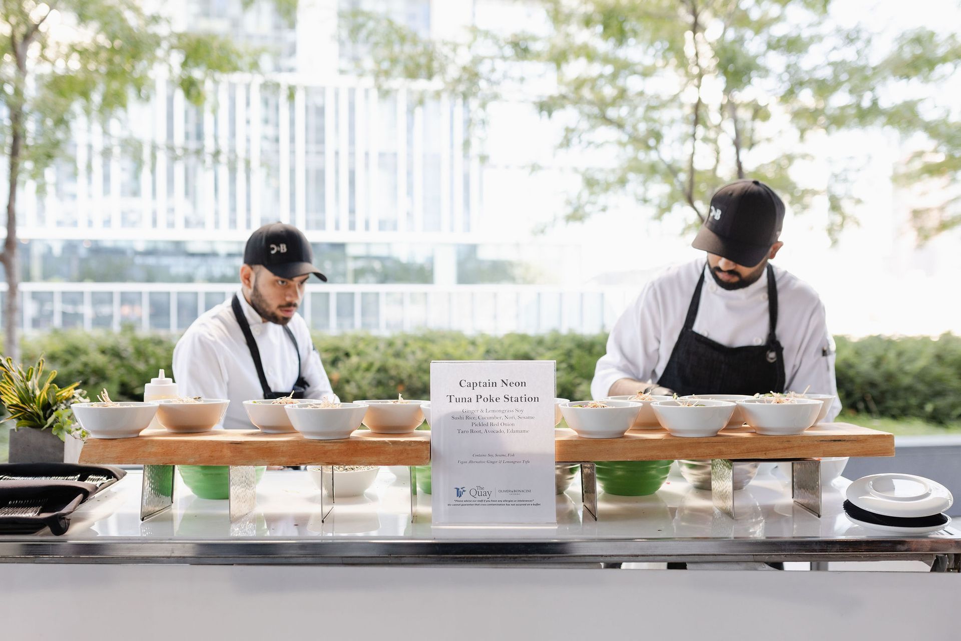 Two chefs prepare food at an outdoor event, serving from bowls on a buffet table.