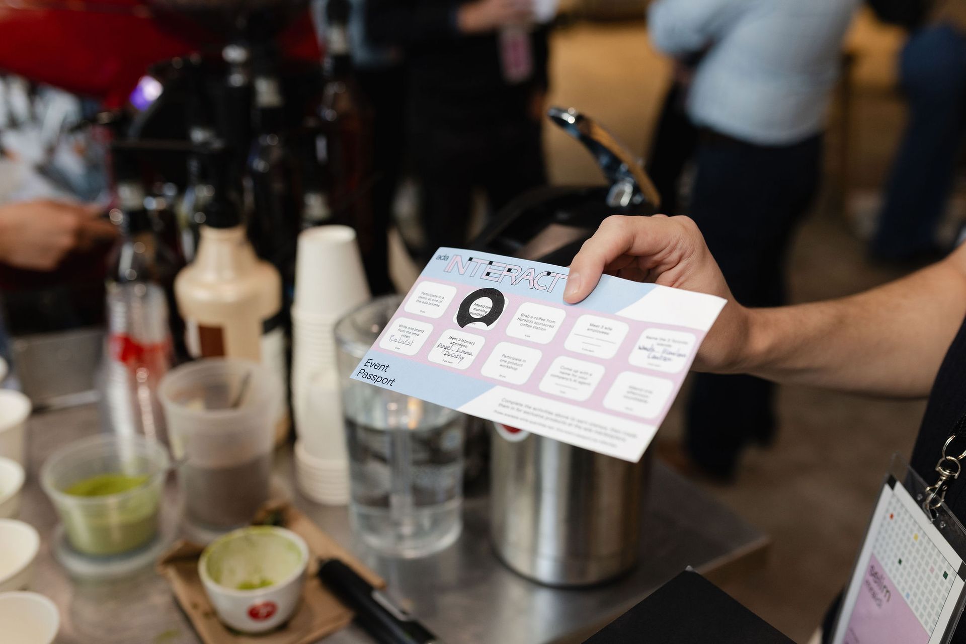 A person holds a promotional card at a cafe counter. Coffee equipment and drinks are nearby.