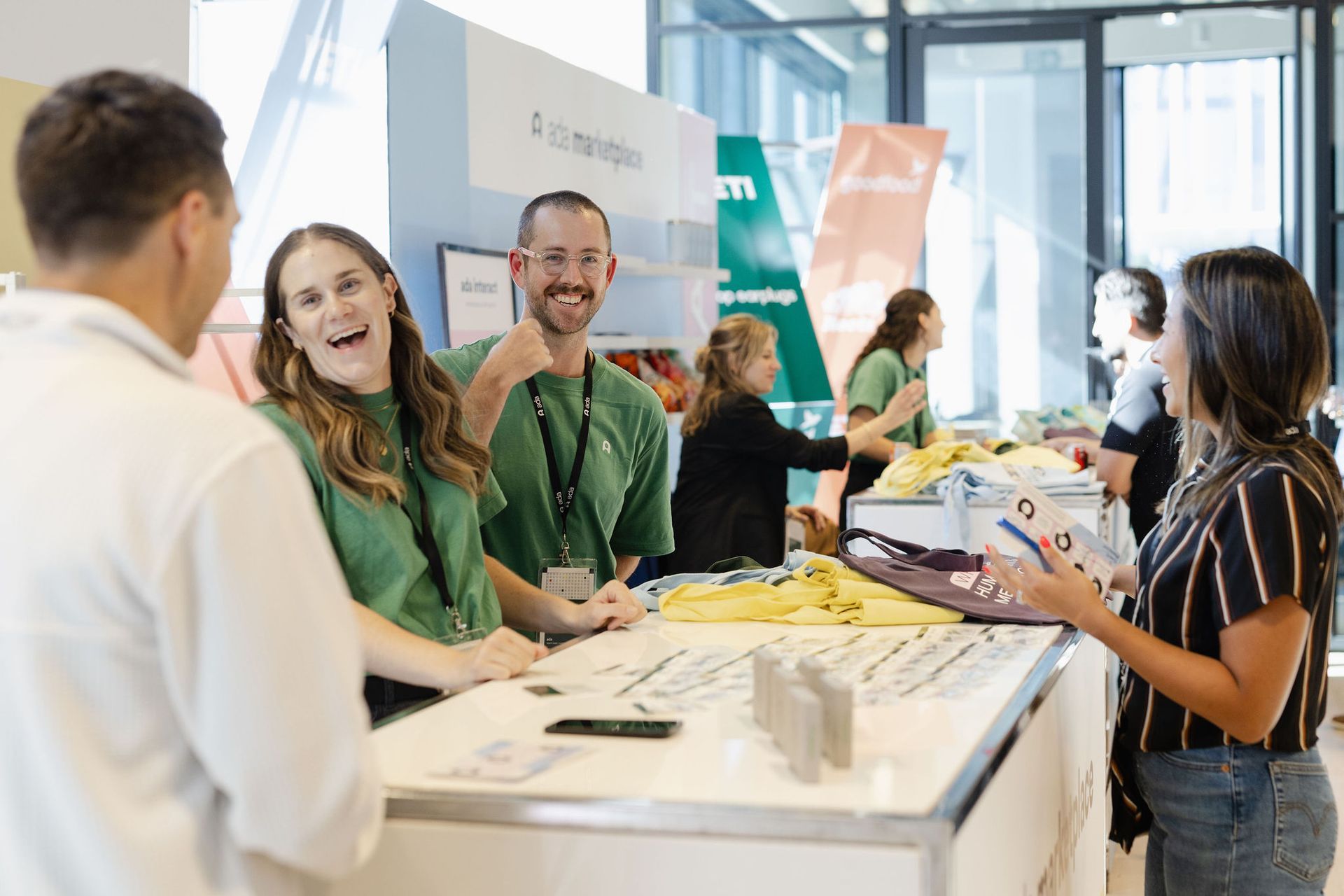 People at an event booth. Smiling people interact at a white table with a green sign and merchandise.
