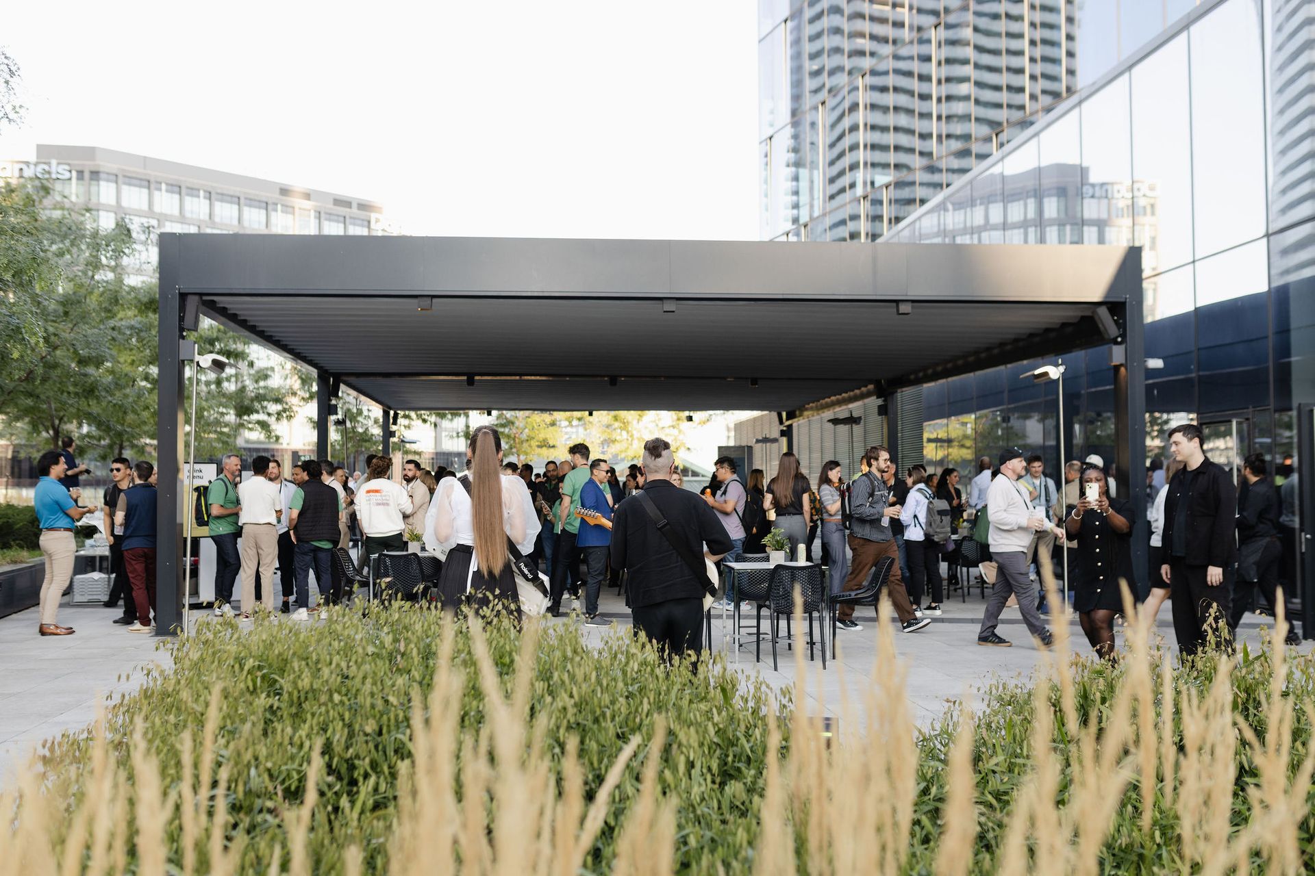 People gathered outdoors under a canopy at an event; modern building in background.