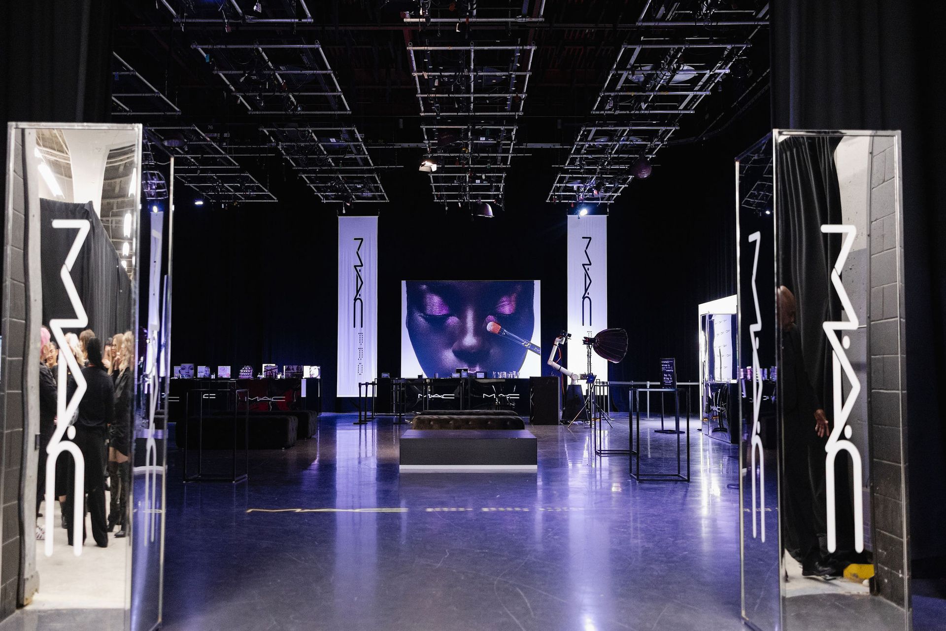 A stage set for a MAC cosmetics event. Two large mirrors frame the entrance. Dark interior, banners, and a central screen.