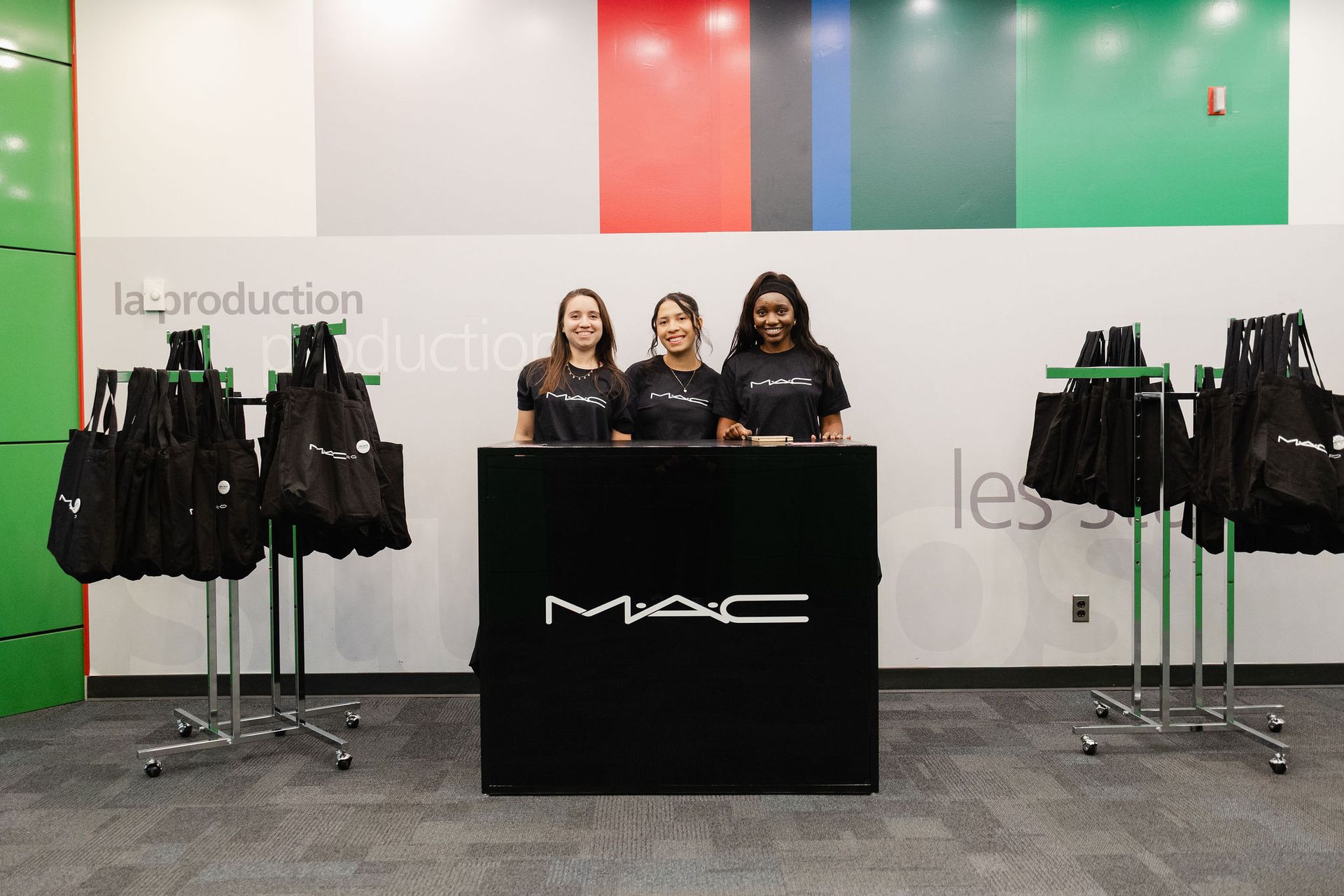 Three people stand behind a MAC cosmetics table with tote bags hanging nearby.