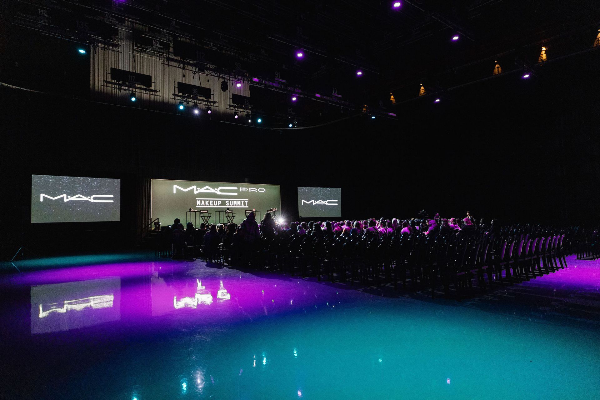 MAC event with a large crowd on a reflective purple and teal floor. MAC logos on screens.