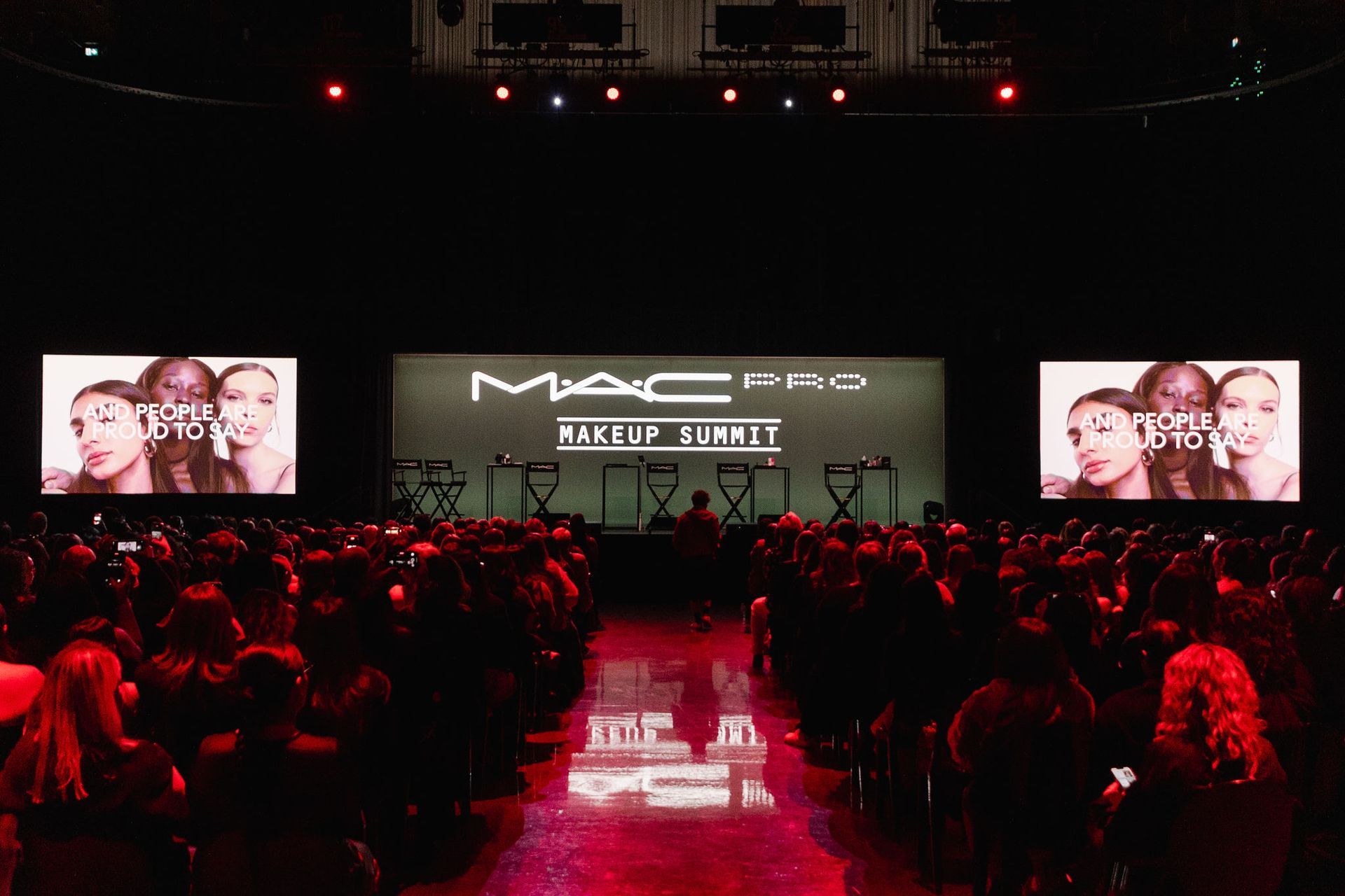MAC Pro makeup event: stage with logo, two screens displaying portraits, audience in red lighting.
