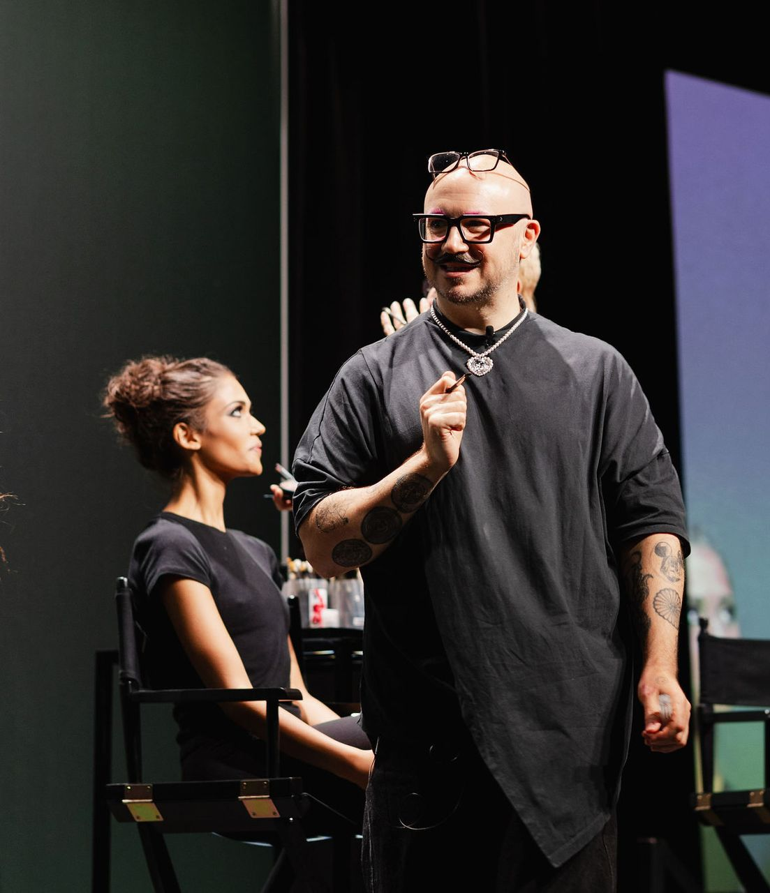 Makeup artist gestures toward model seated in a chair, both on a stage.
