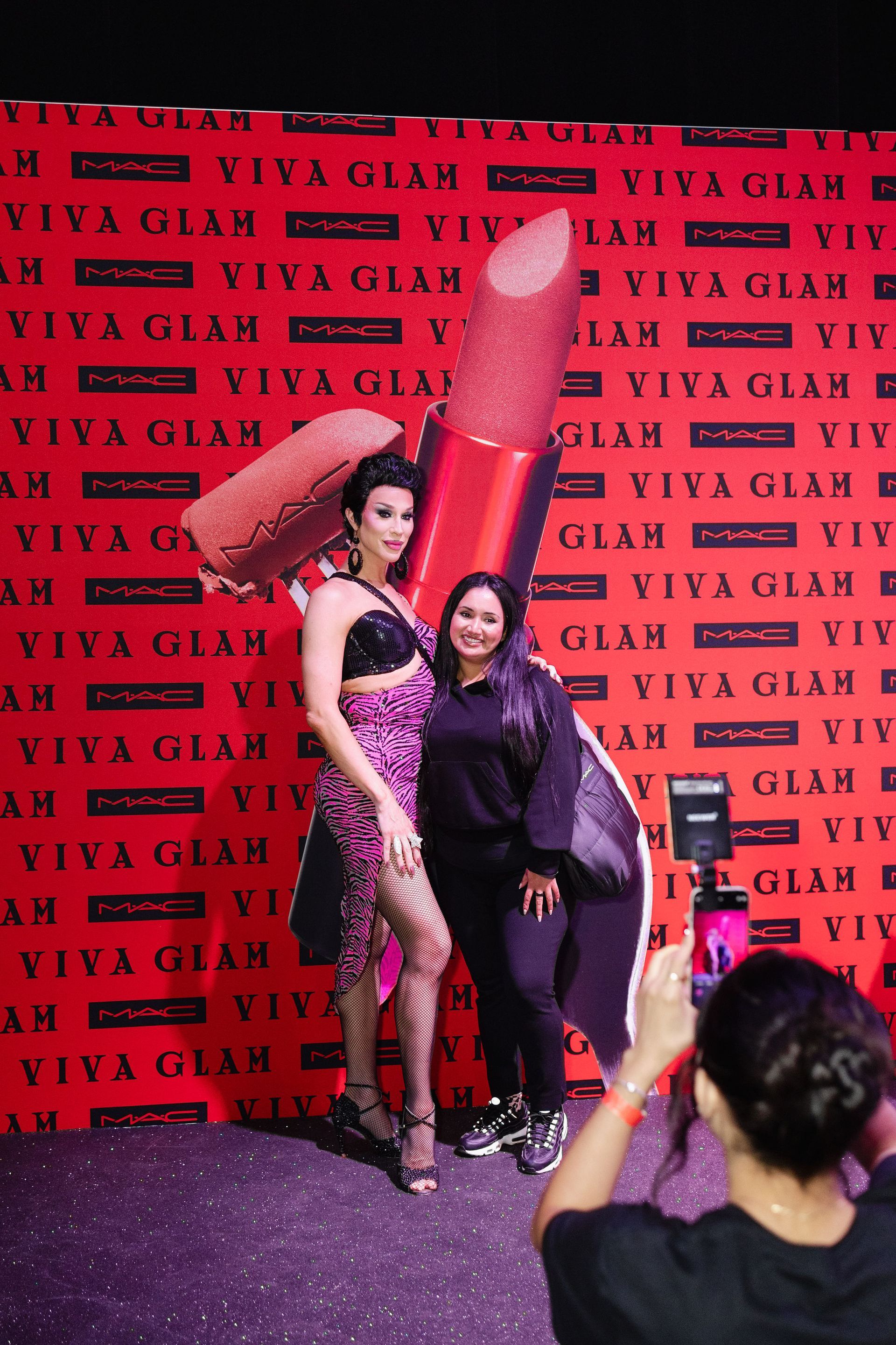 Person posing with a drag queen in front of a 