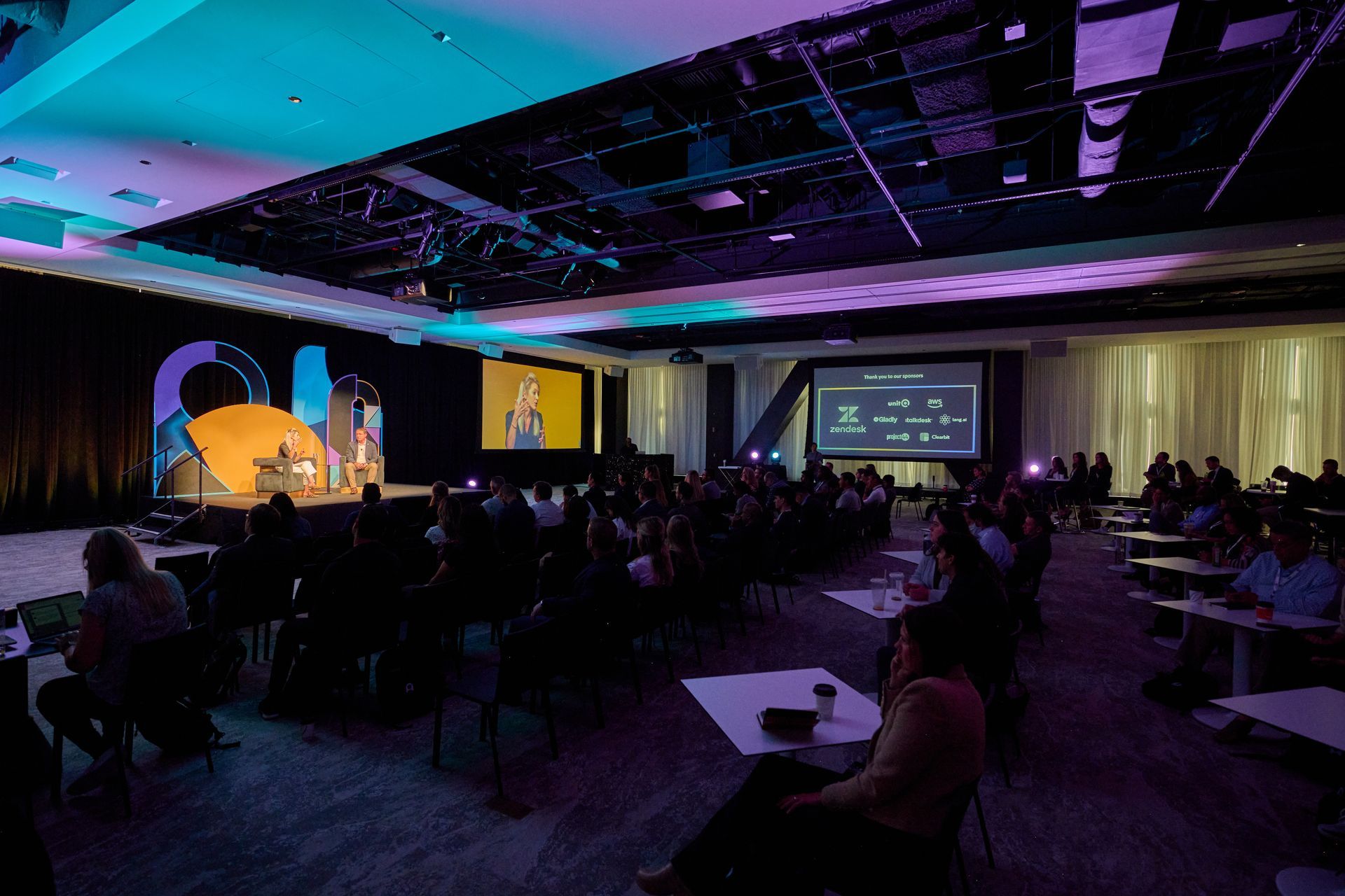 A large room filled with people sitting at tables and watching a presentation.