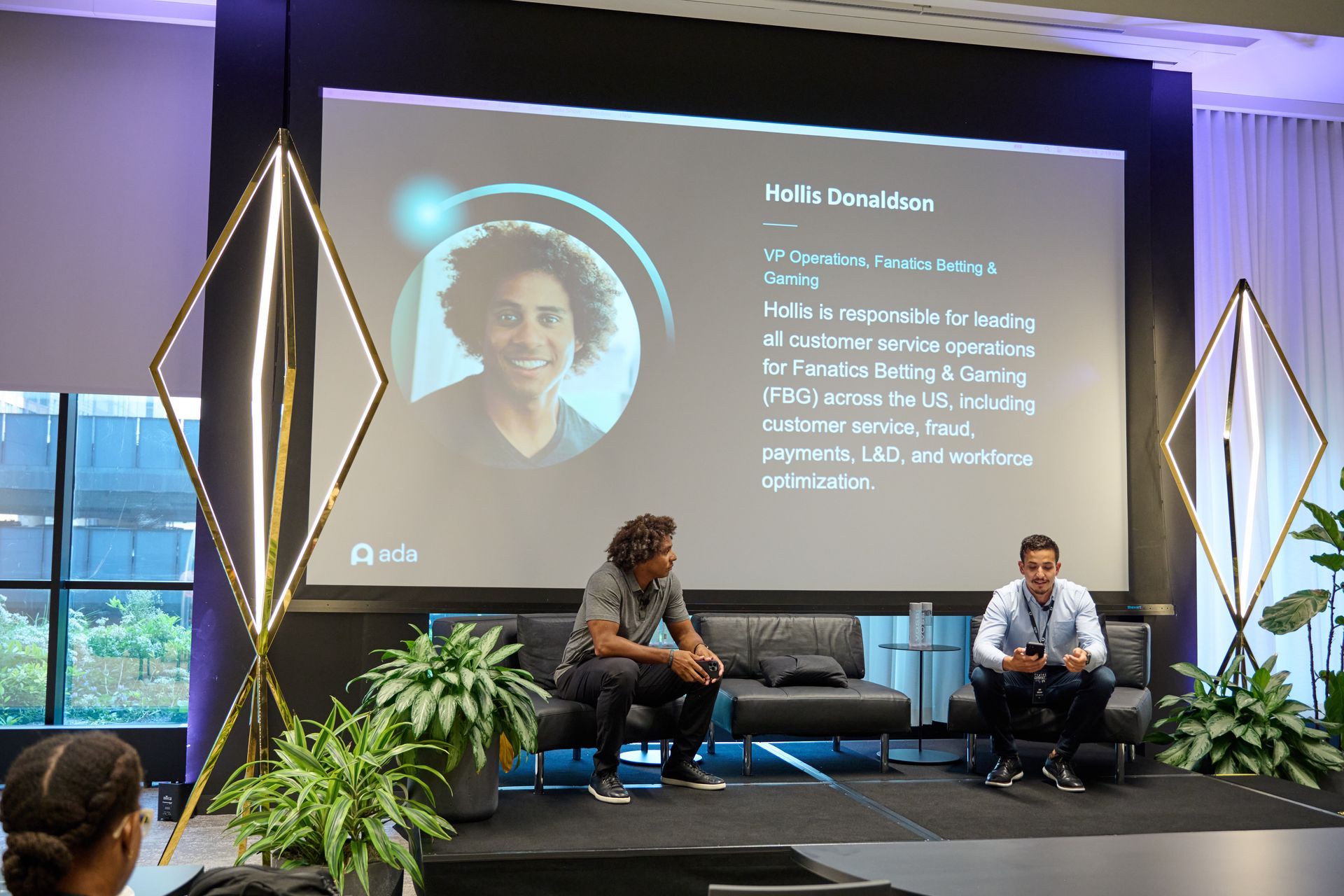 Two men are sitting in front of a large screen with a picture of a man on it.