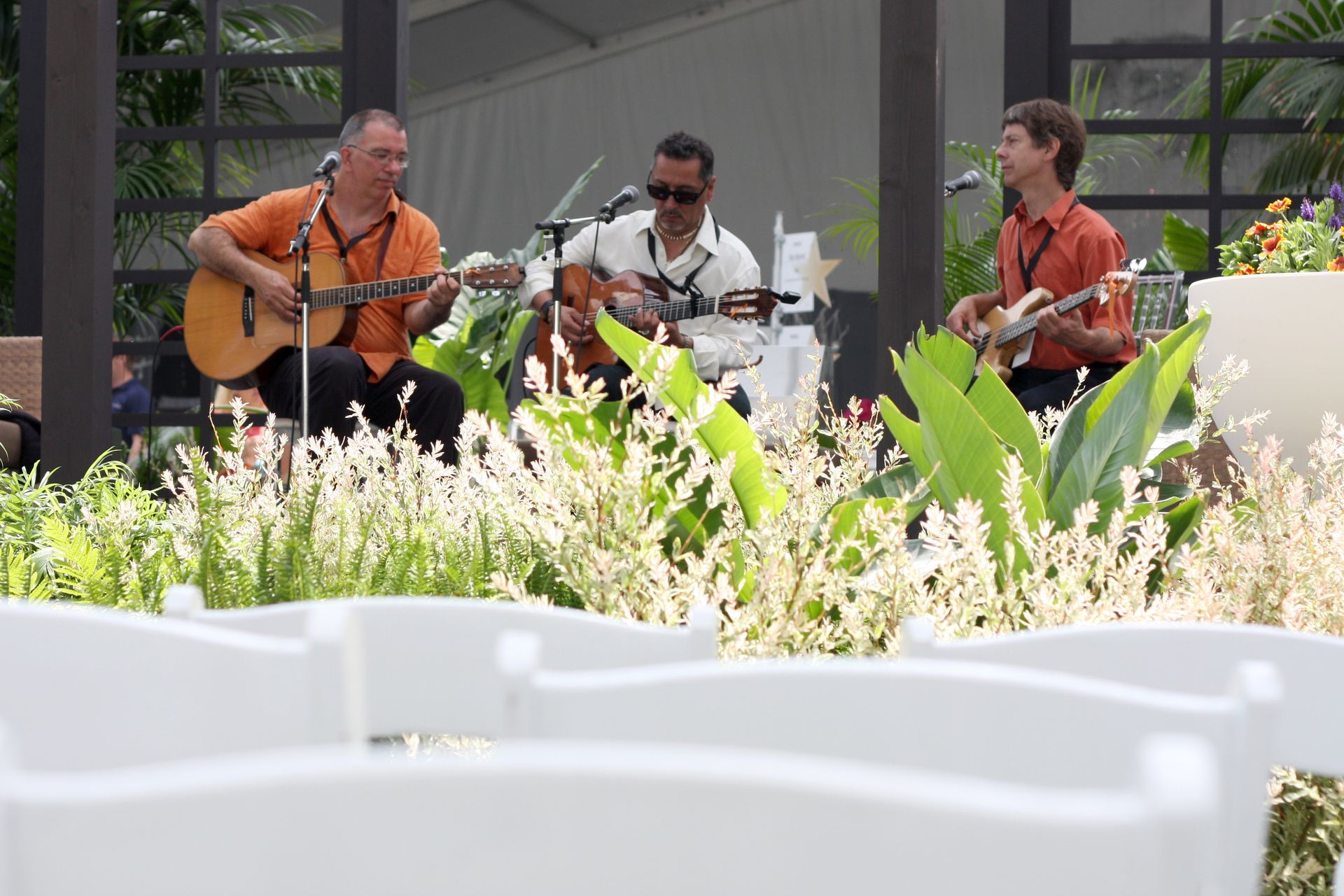 Three musicians play guitars outdoors; white chairs in foreground; lush greenery surrounds the stage.