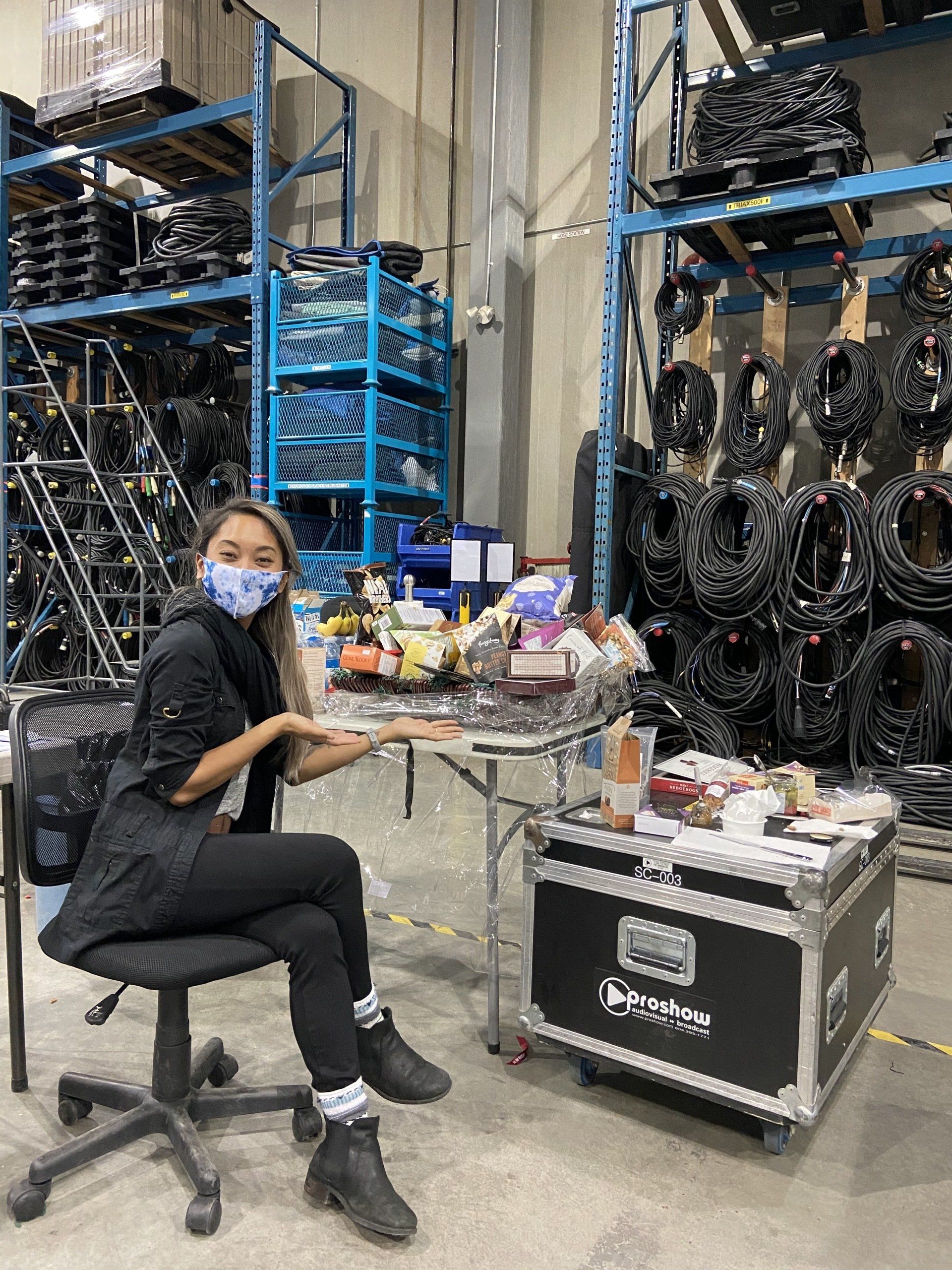 A woman wearing a mask is sitting at a table in a warehouse.