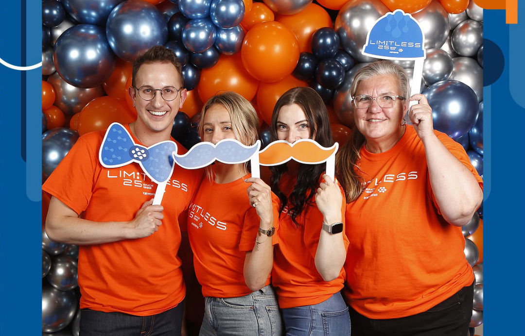 A group of people standing in front of a wall of balloons holding fake mustaches and bow ties.