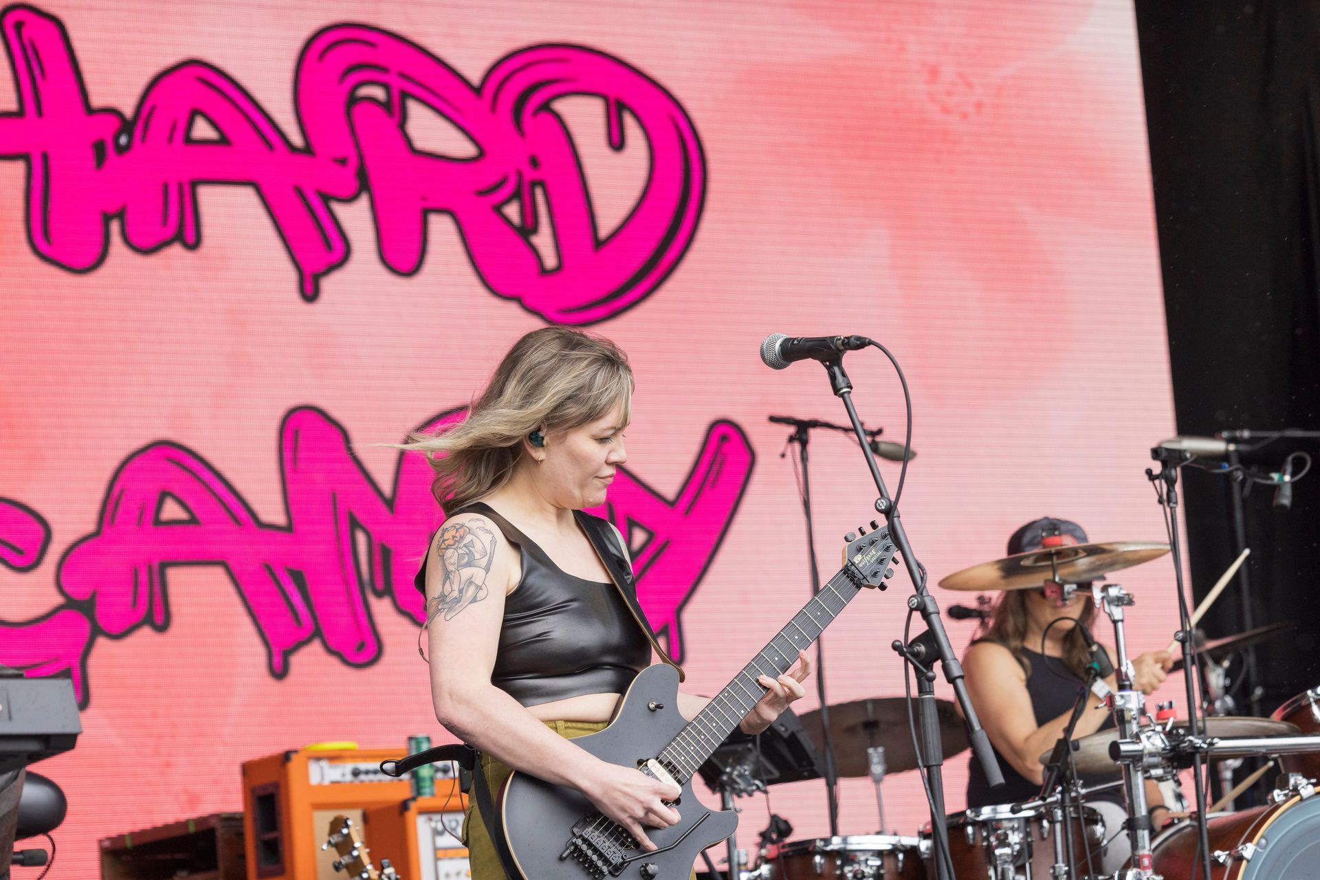 Woman playing electric guitar onstage with drummer behind her; pink 