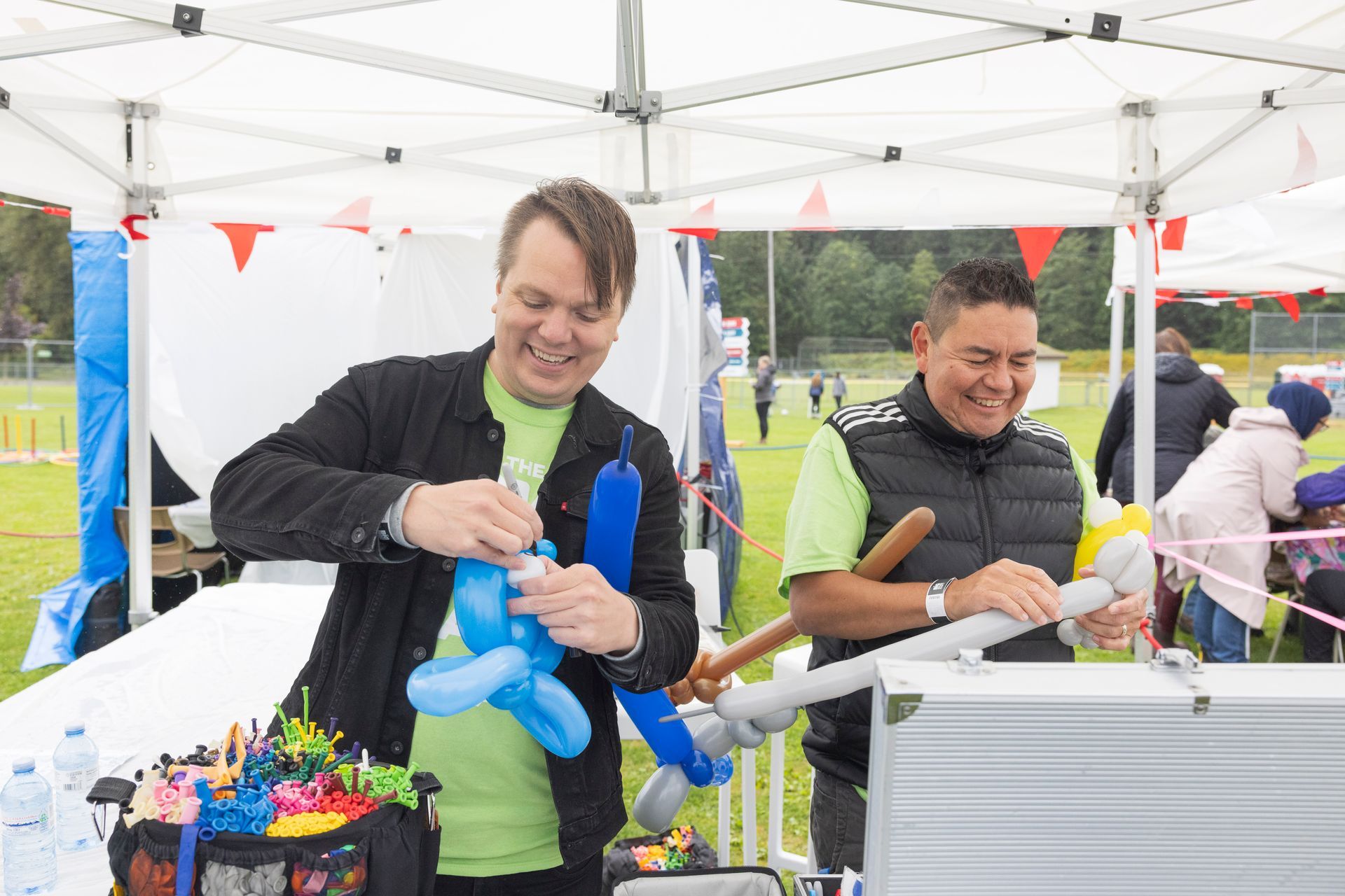 Two men smiling, twisting balloons under a white tent at an outdoor event.