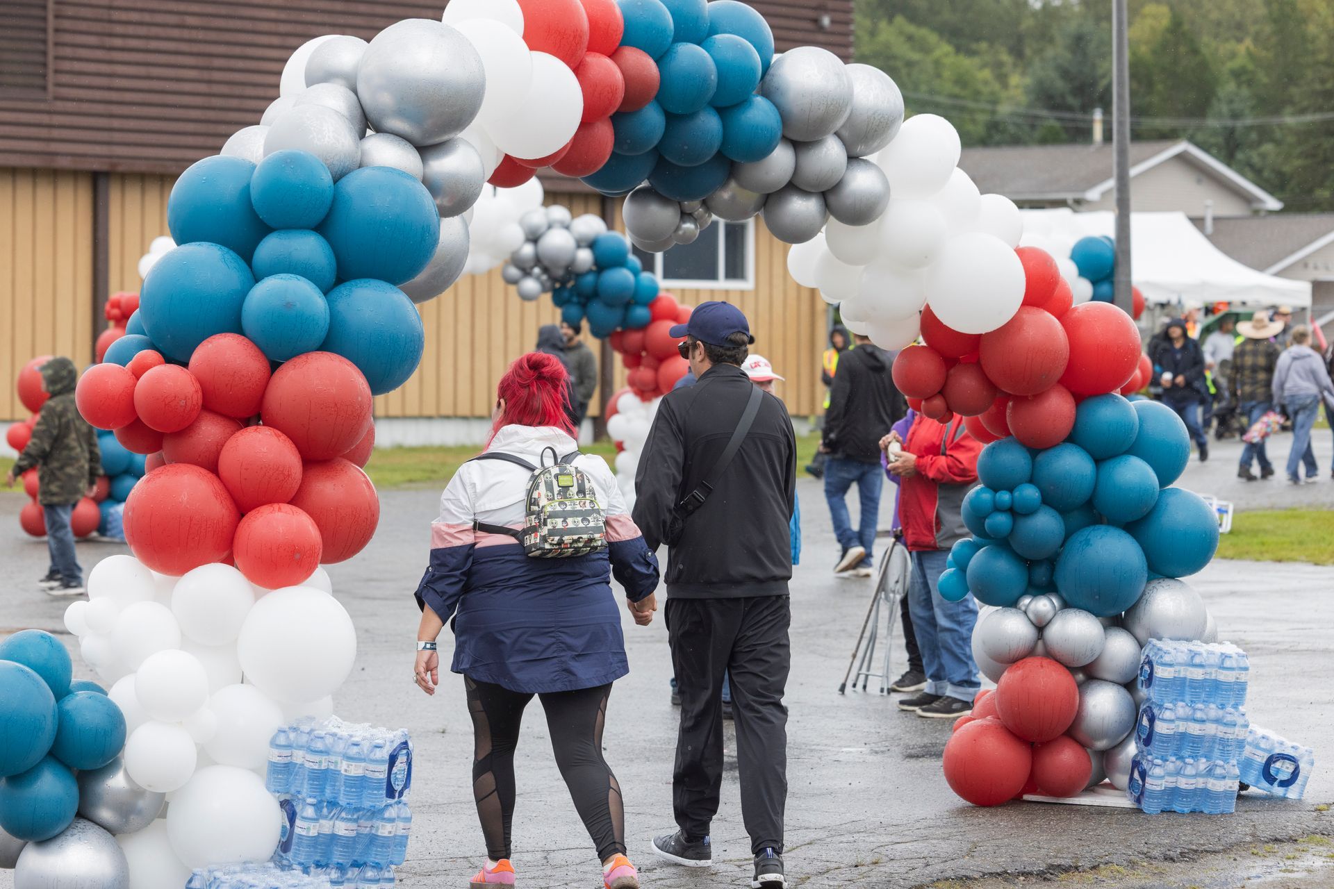 People walk under a red, white, and blue balloon arch at an outdoor event.
