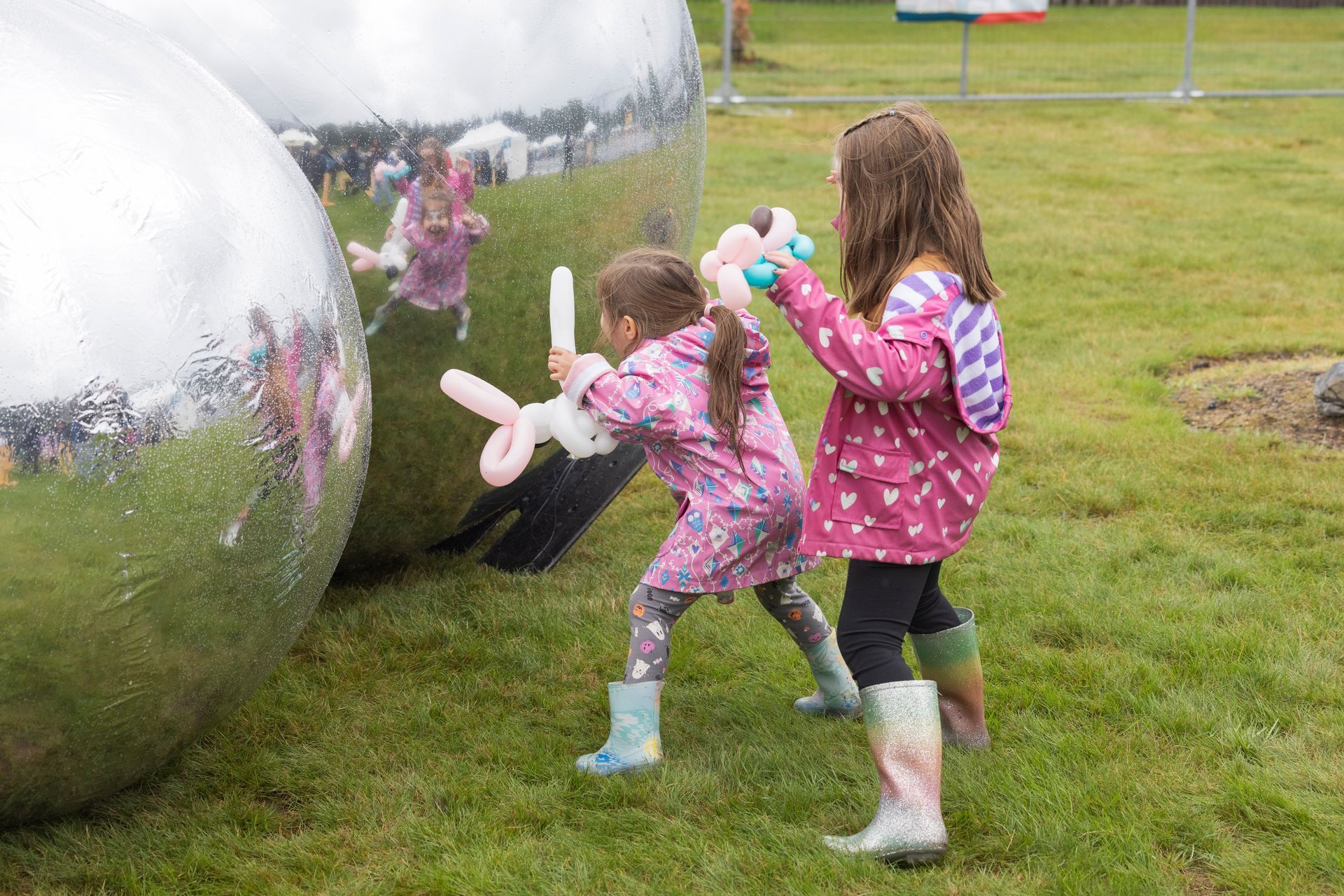 Two children in raincoats play with balloon animals near large reflective spheres on grass.