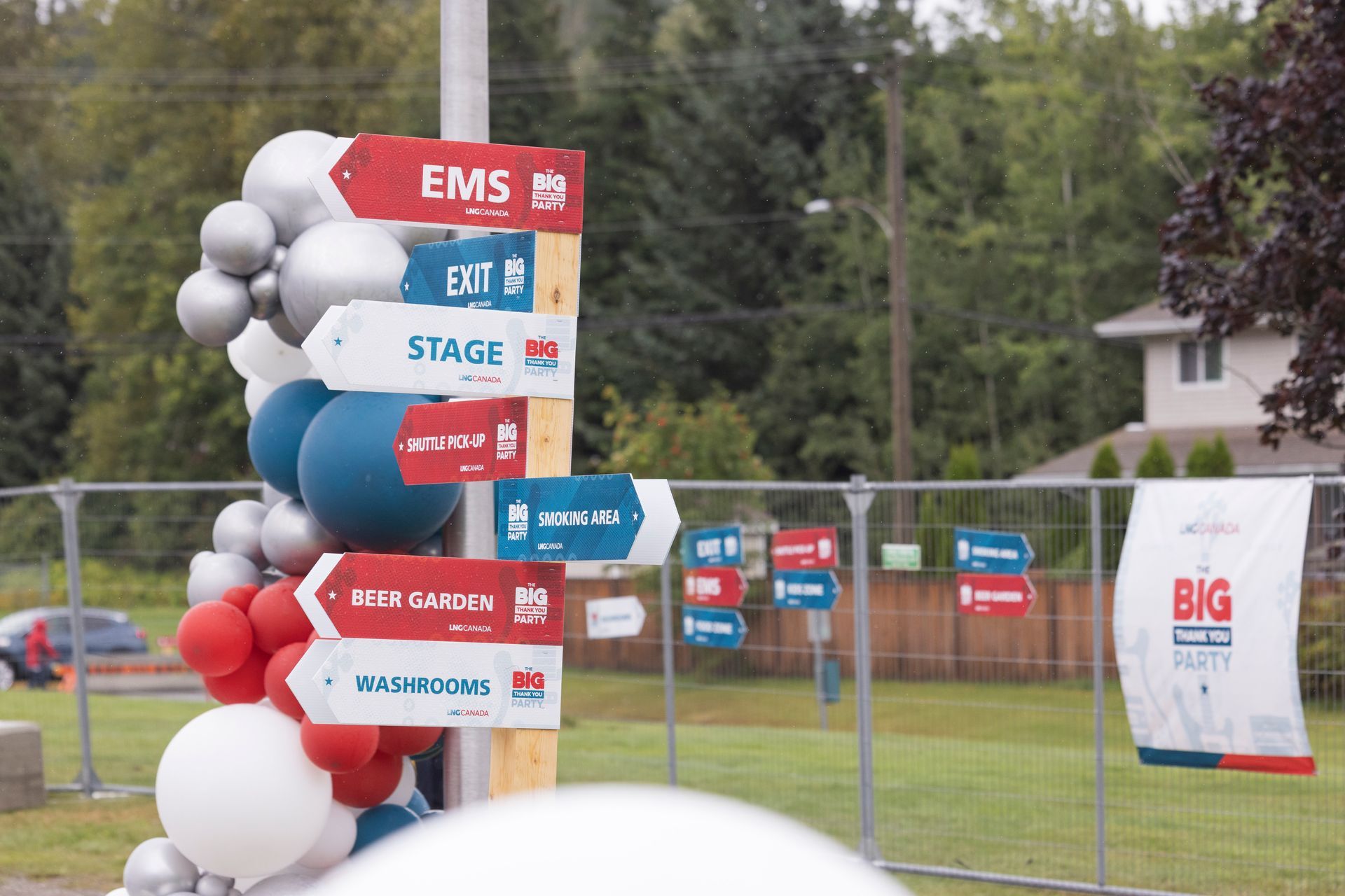 Directional signs at an outdoor event, listing destinations. Balloons in red, white, and blue decorate the pole.
