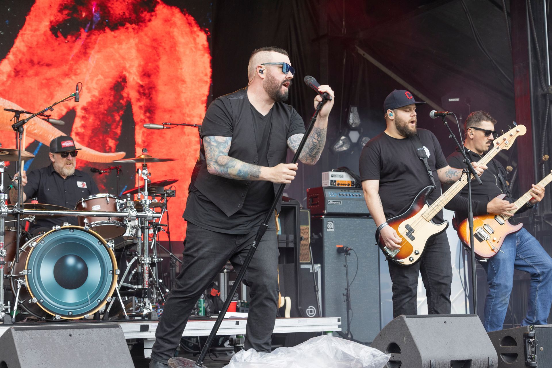 Band performing on stage, singer with tattoos, drums, bass guitars, large red backdrop.
