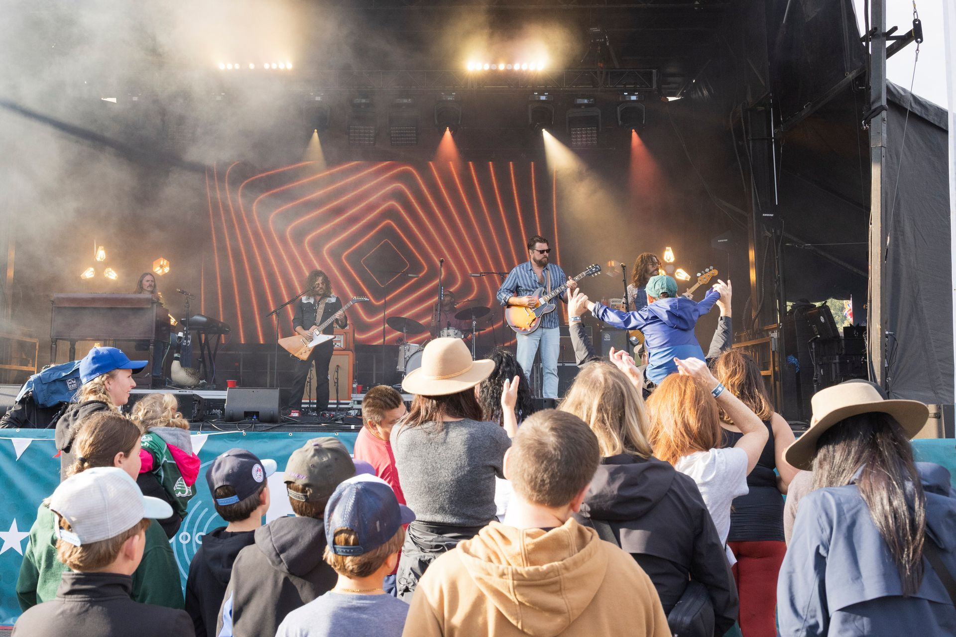 Band performs on stage at outdoor festival, crowd watches. Stage lit with geometric lights and fog.