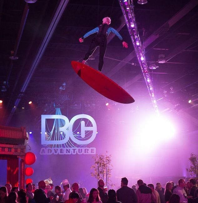 A man is flying through the air on a surfboard in front of a sign that says vbg adventure