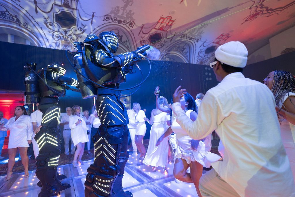 Two robot performers with lights dance at a party with people in white clothing.