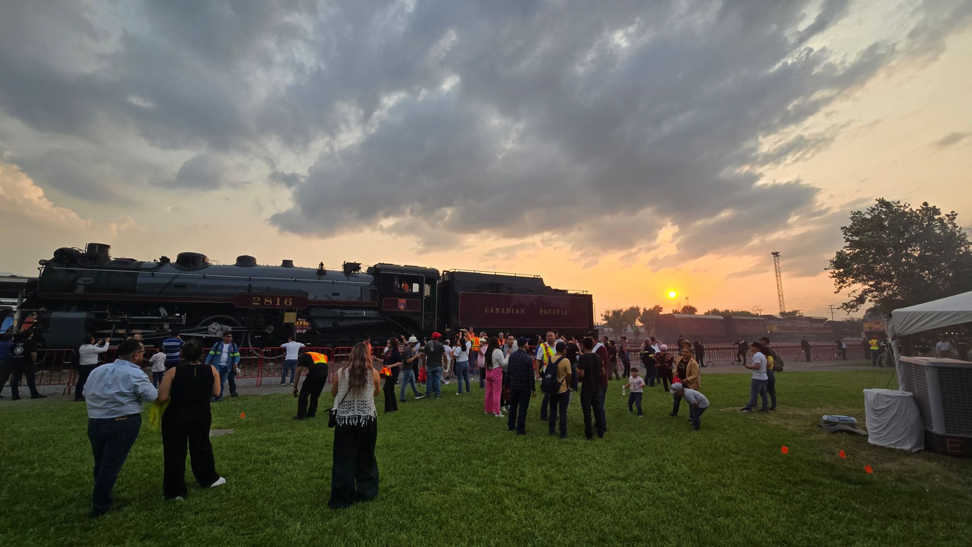 A group of people are standing in a field in front of a train at sunset.