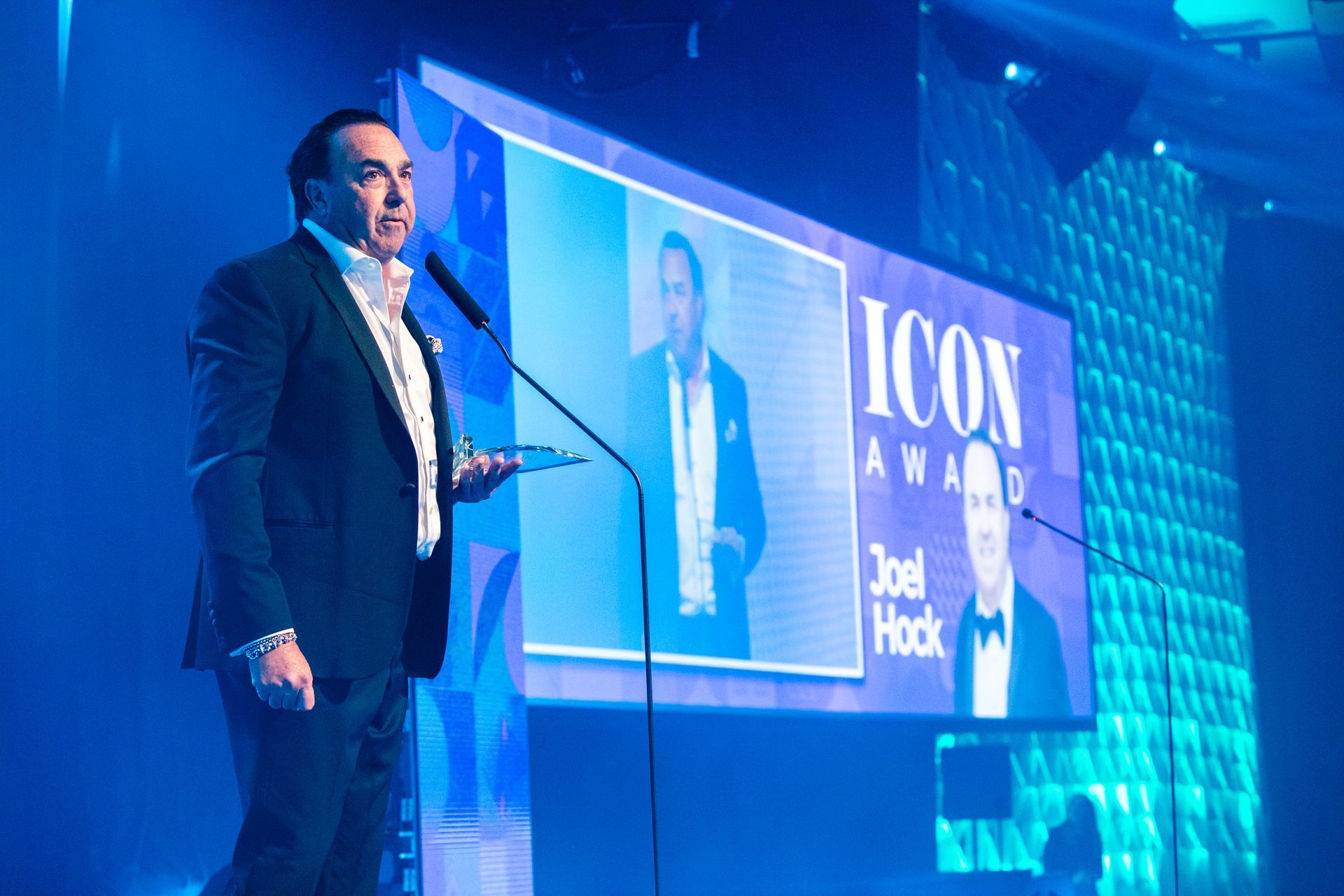 Man holding award, speaking at podium with screen behind him, lit with blue light, saying “ICON AWARD Joel Hock”.