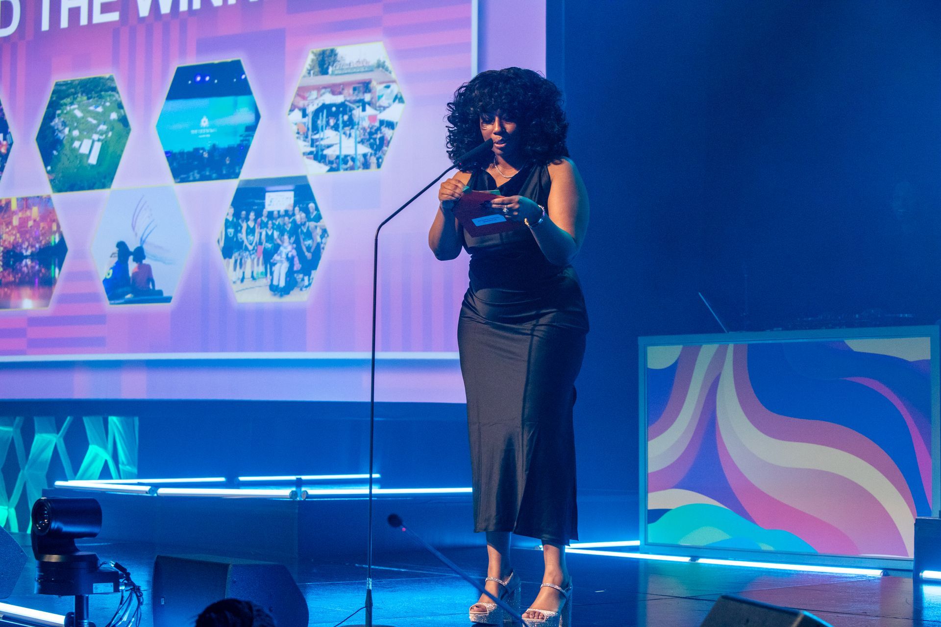 Woman in black dress speaks at podium on stage with screen displaying images.