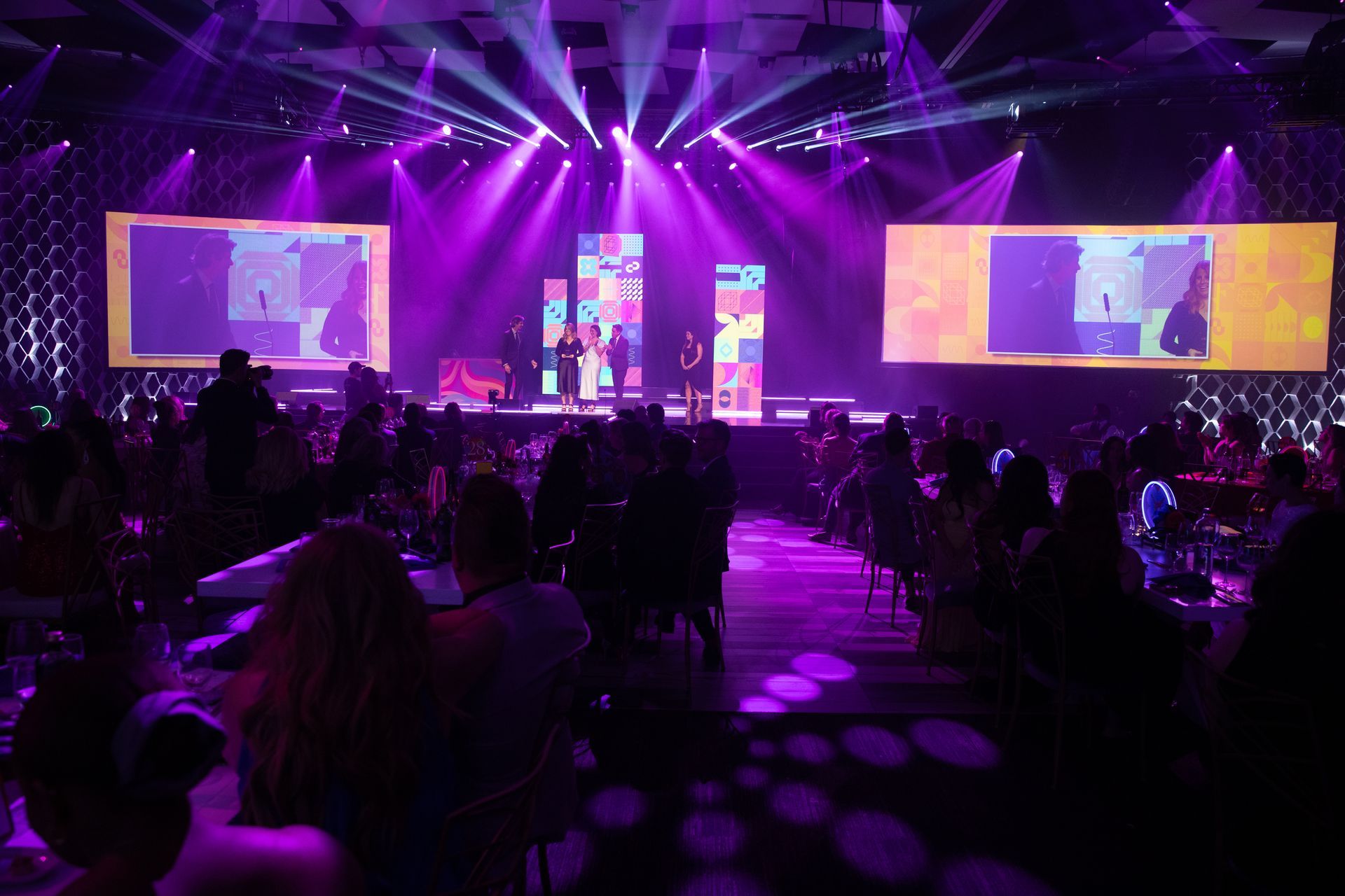 Purple-lit stage with performers, screens displaying images, and an audience seated at tables.