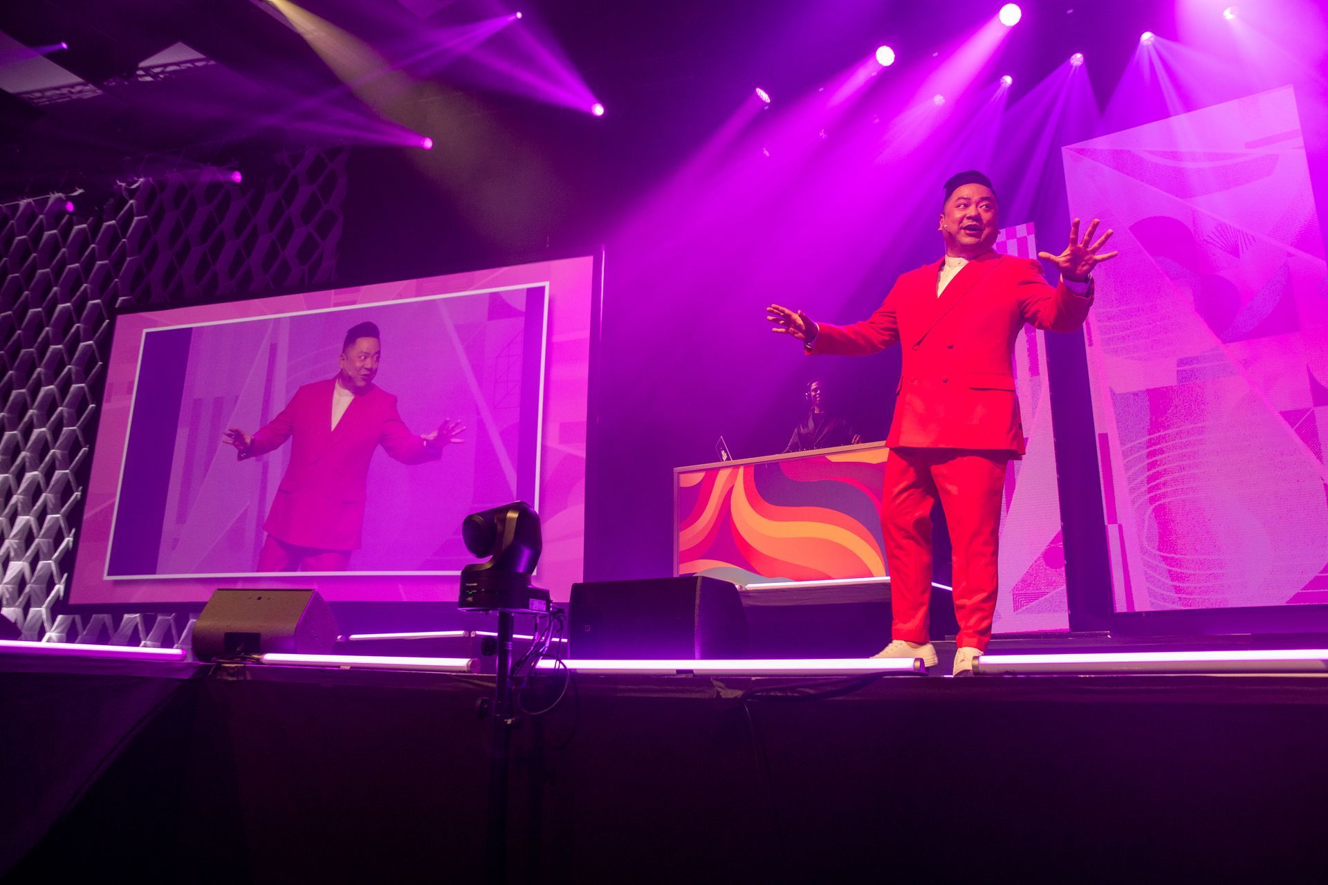 Man in a red suit on a stage, performing with a screen showing his image and bright pink lighting.