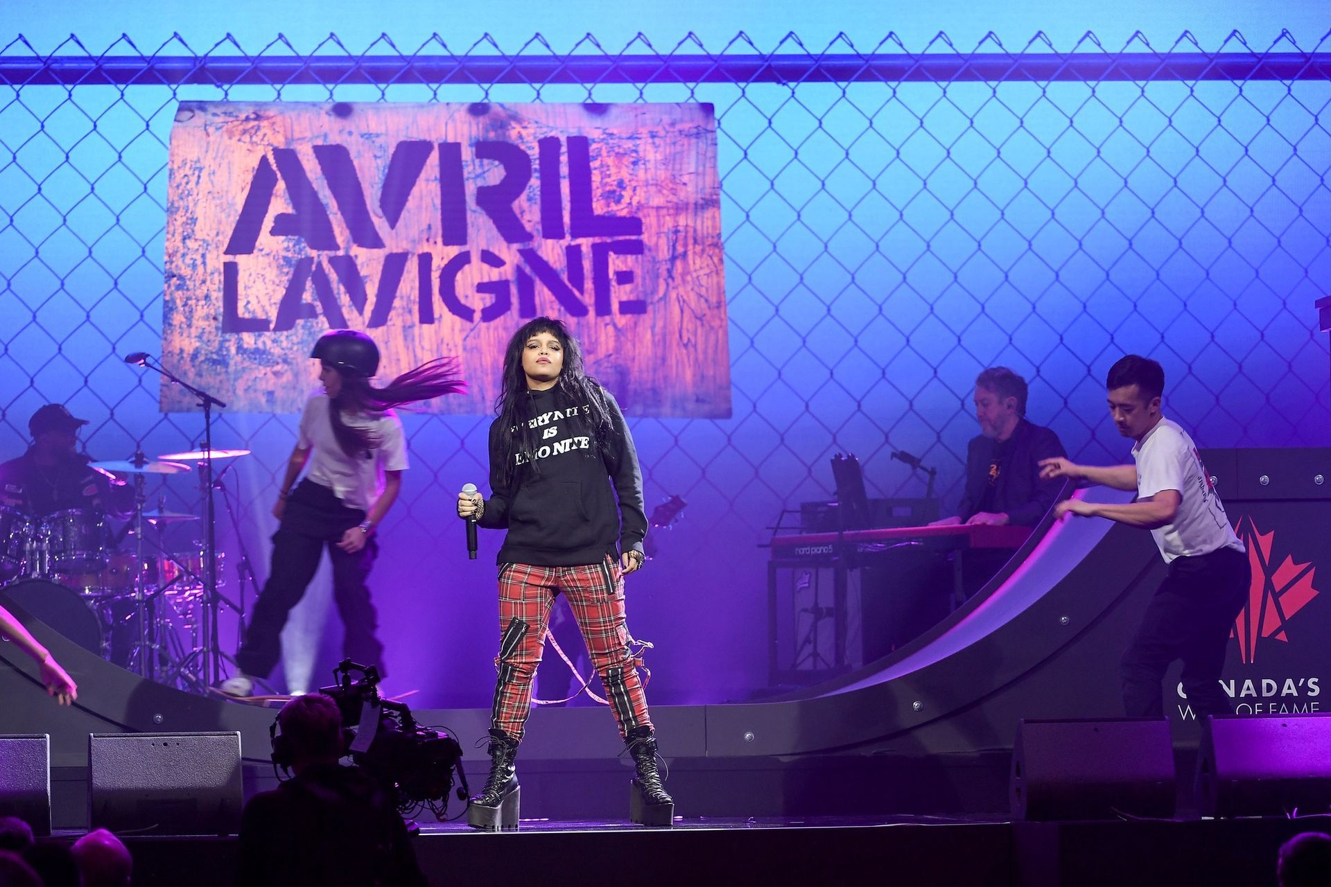 Avril Lavigne performing on stage with band, wearing hoodie and patterned pants; stage backdrop with her name.