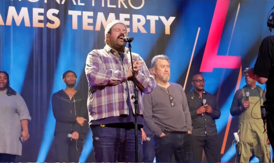 A man singing into a microphone on a stage in front of a sign that says mes temerty