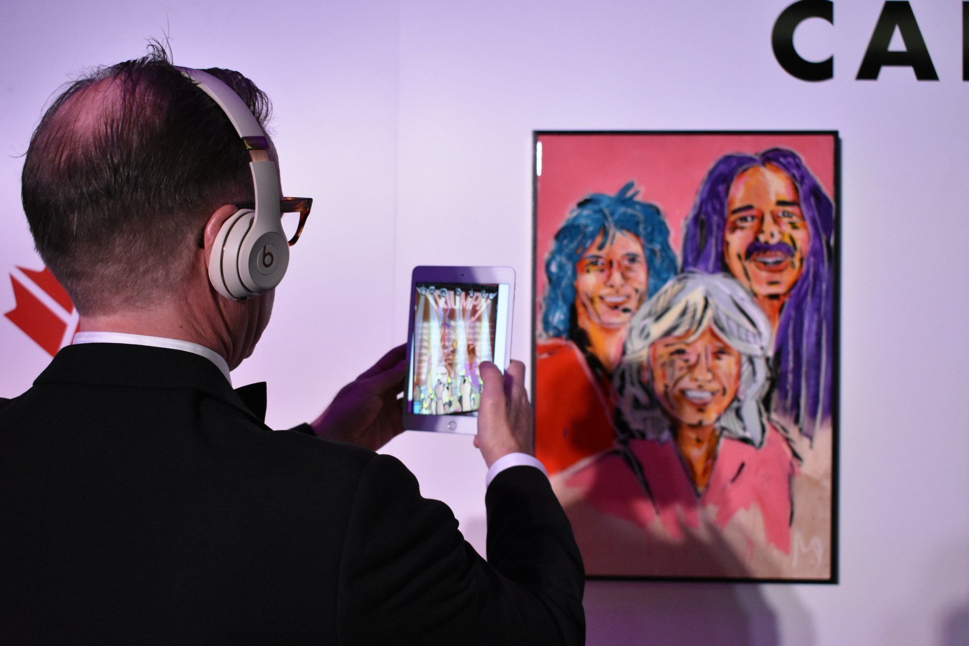 A man wearing headphones is taking a picture of a painting on a wall.