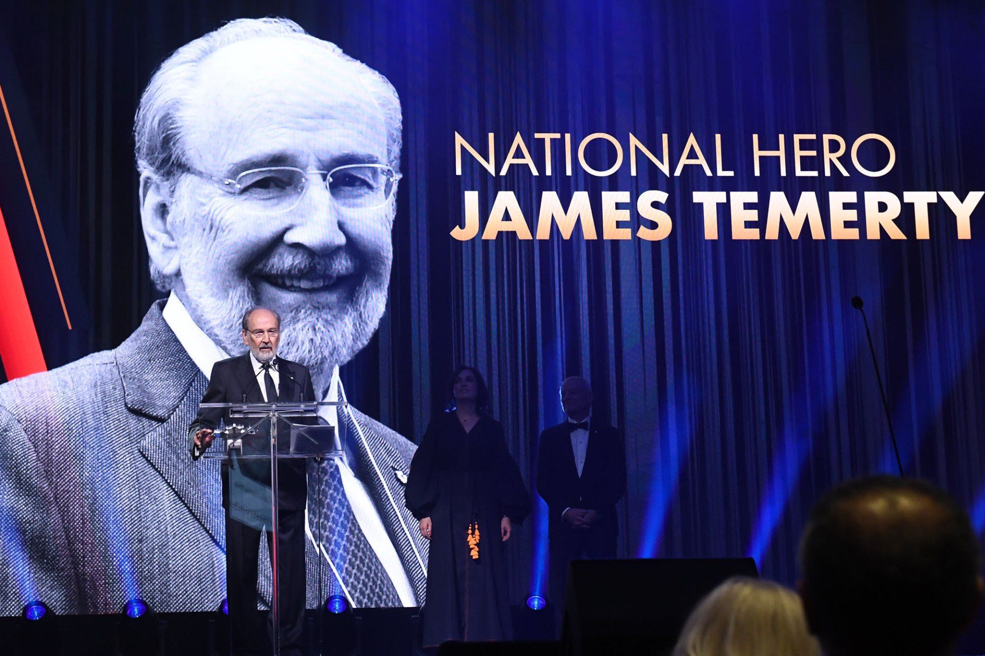 A man is giving a speech in front of a screen that says national hero james temerty