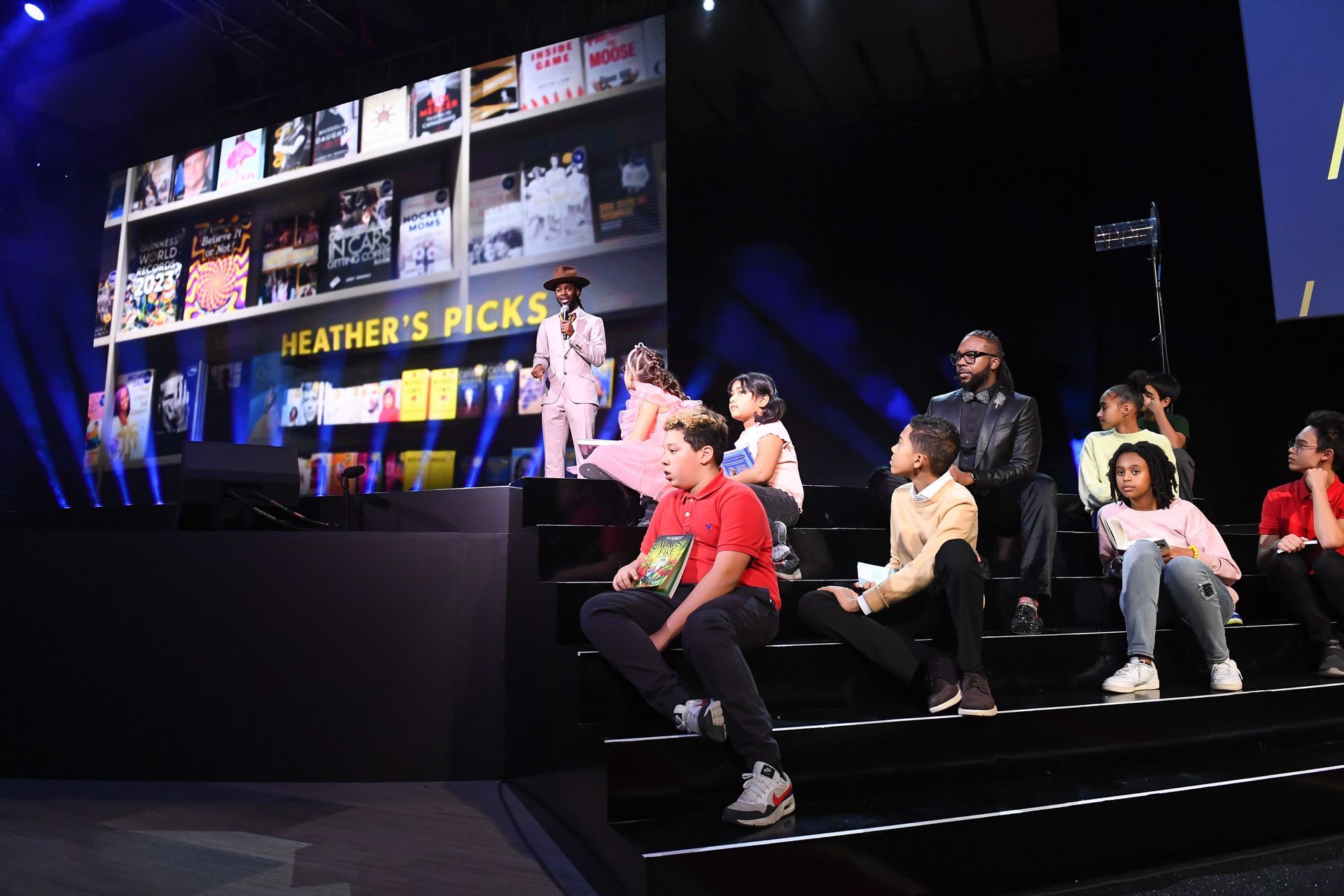 A group of people are sitting on a stage in front of a large screen that says heather 's picks.