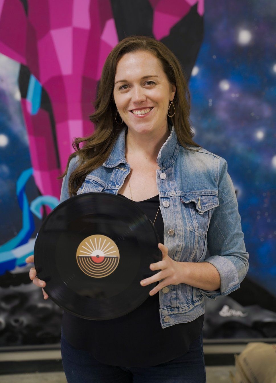 A woman in a denim jacket is holding a record and smiling.