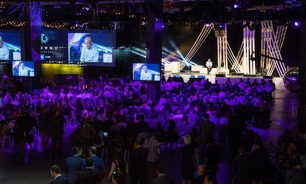 A large crowd of people are gathered in a large room with purple lights.