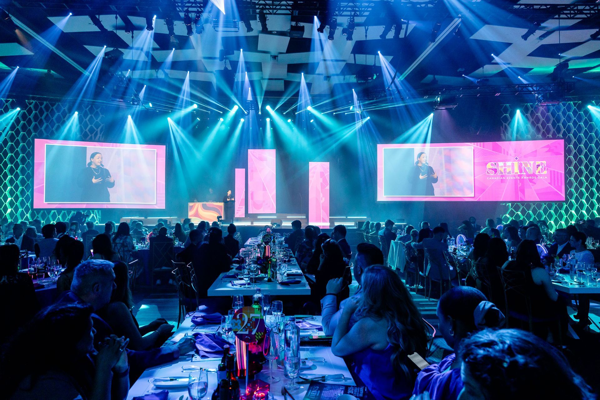 A brightly lit awards ceremony with stage, screens, audience at tables, and spotlights shining down.