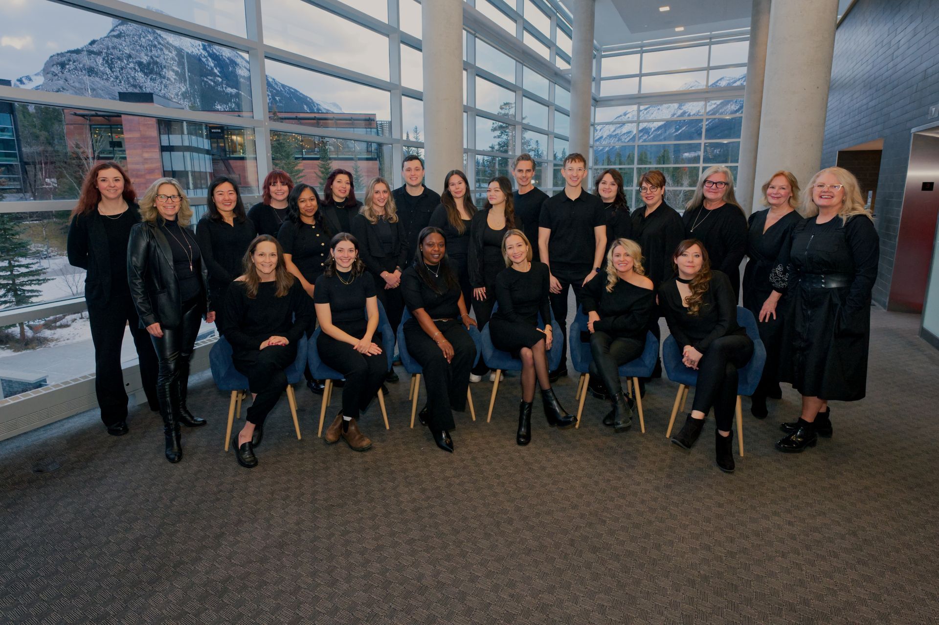 Group of people in black attire posing indoors, windows and mountains in the background.