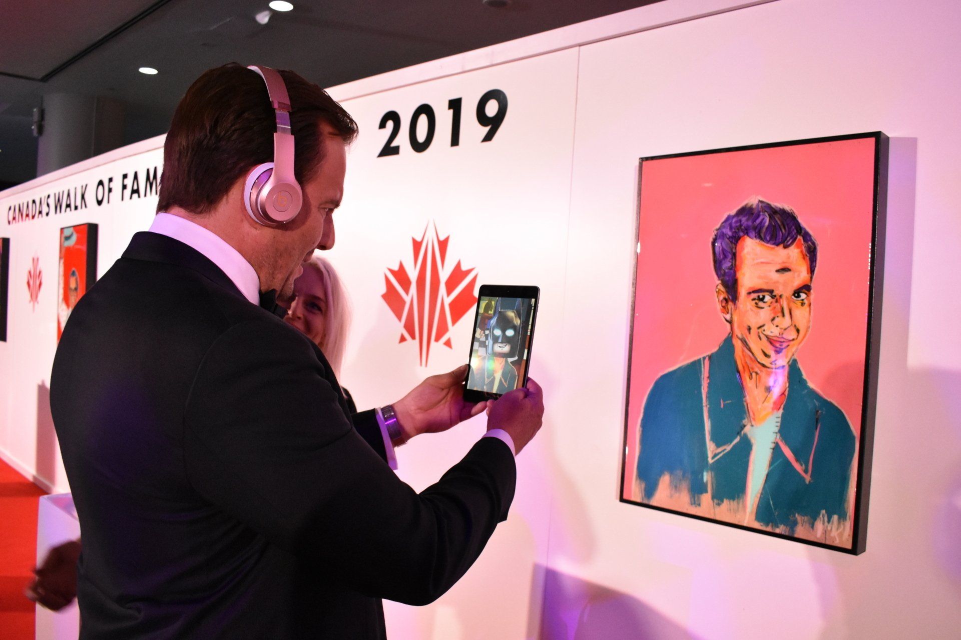 A man wearing headphones is taking a picture of a painting on a wall.