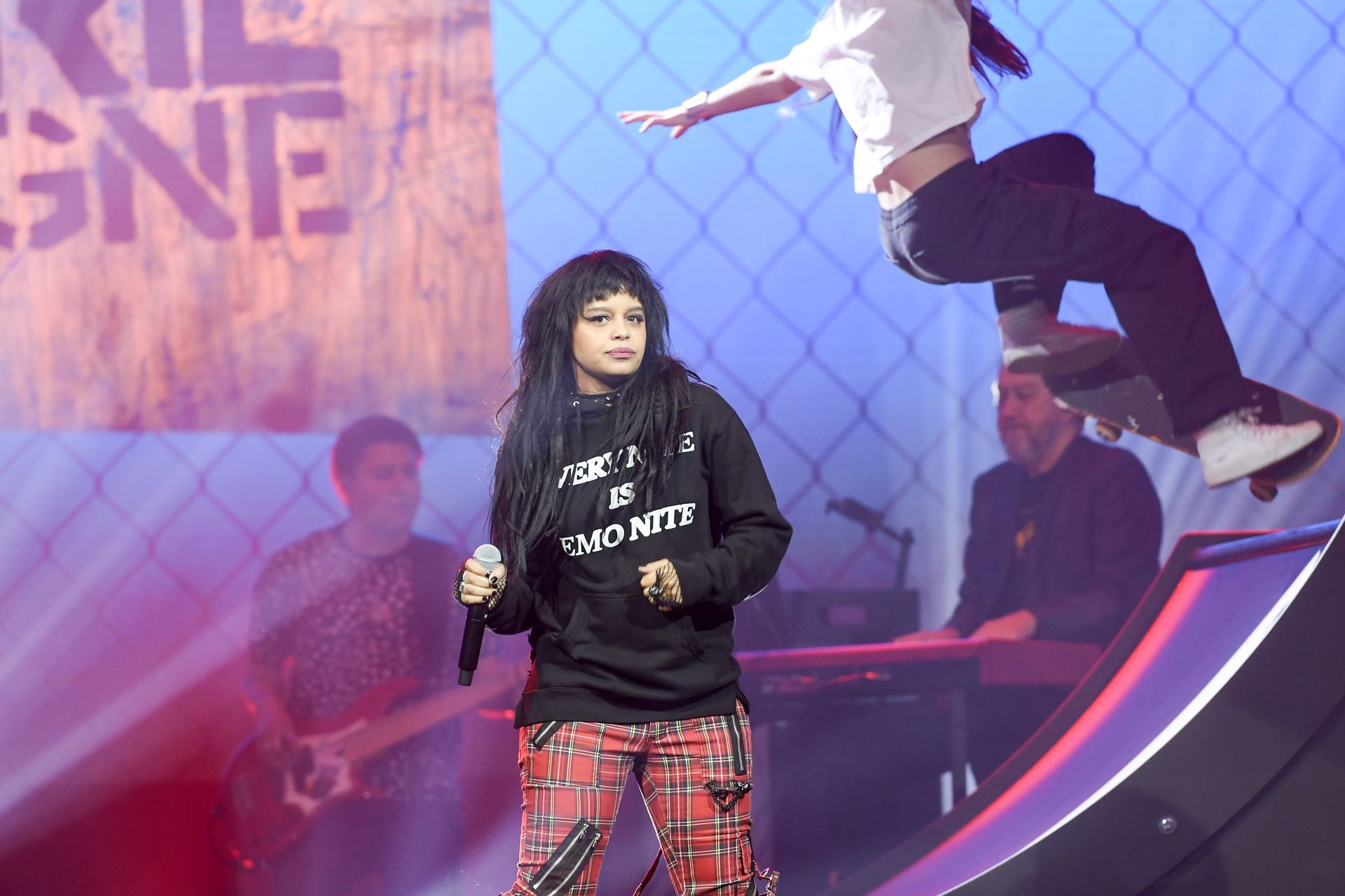 A woman is singing into a microphone while a skateboarder is jumping in the air behind her.