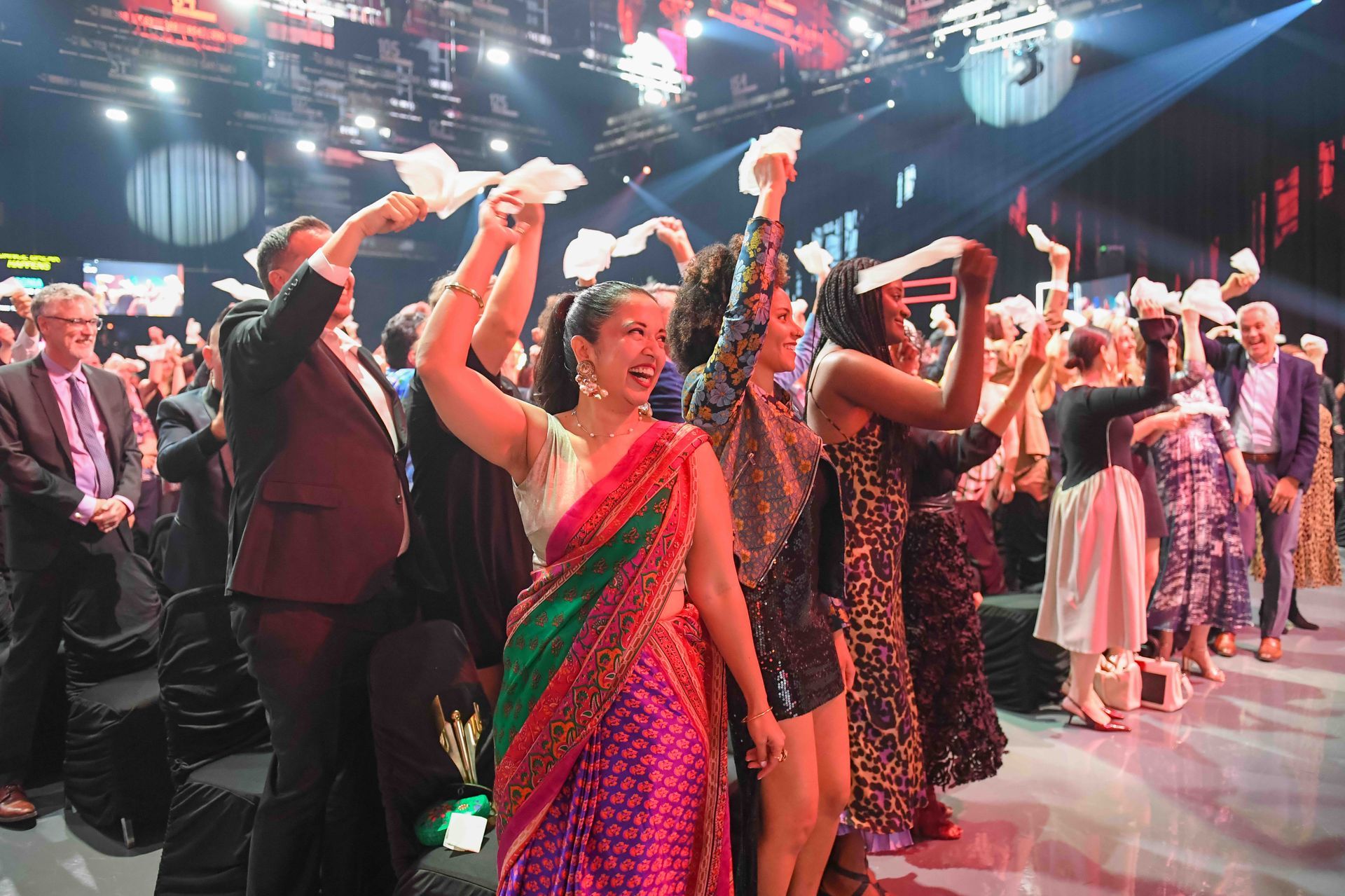 People in formal wear holding up papers, celebrating in a brightly lit auditorium.
