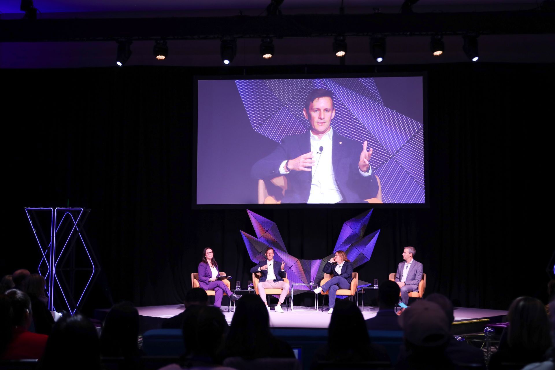 Panel discussion on stage with speaker on screen. Purple and blue lighting. Audience seated.
