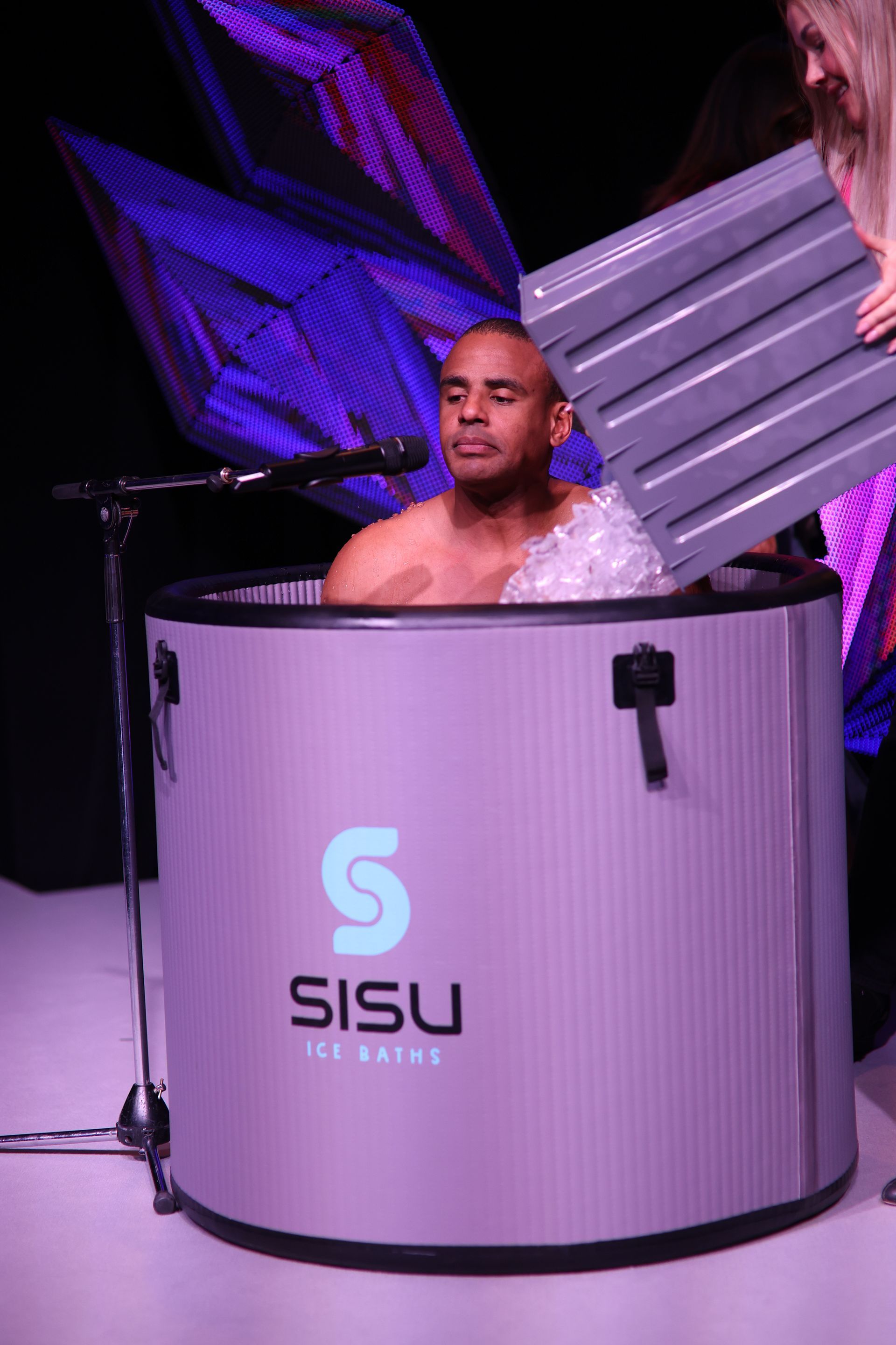 Man in a SISU ice bath. Woman places cover over the bath. Purple tub with logo. Stage setting.