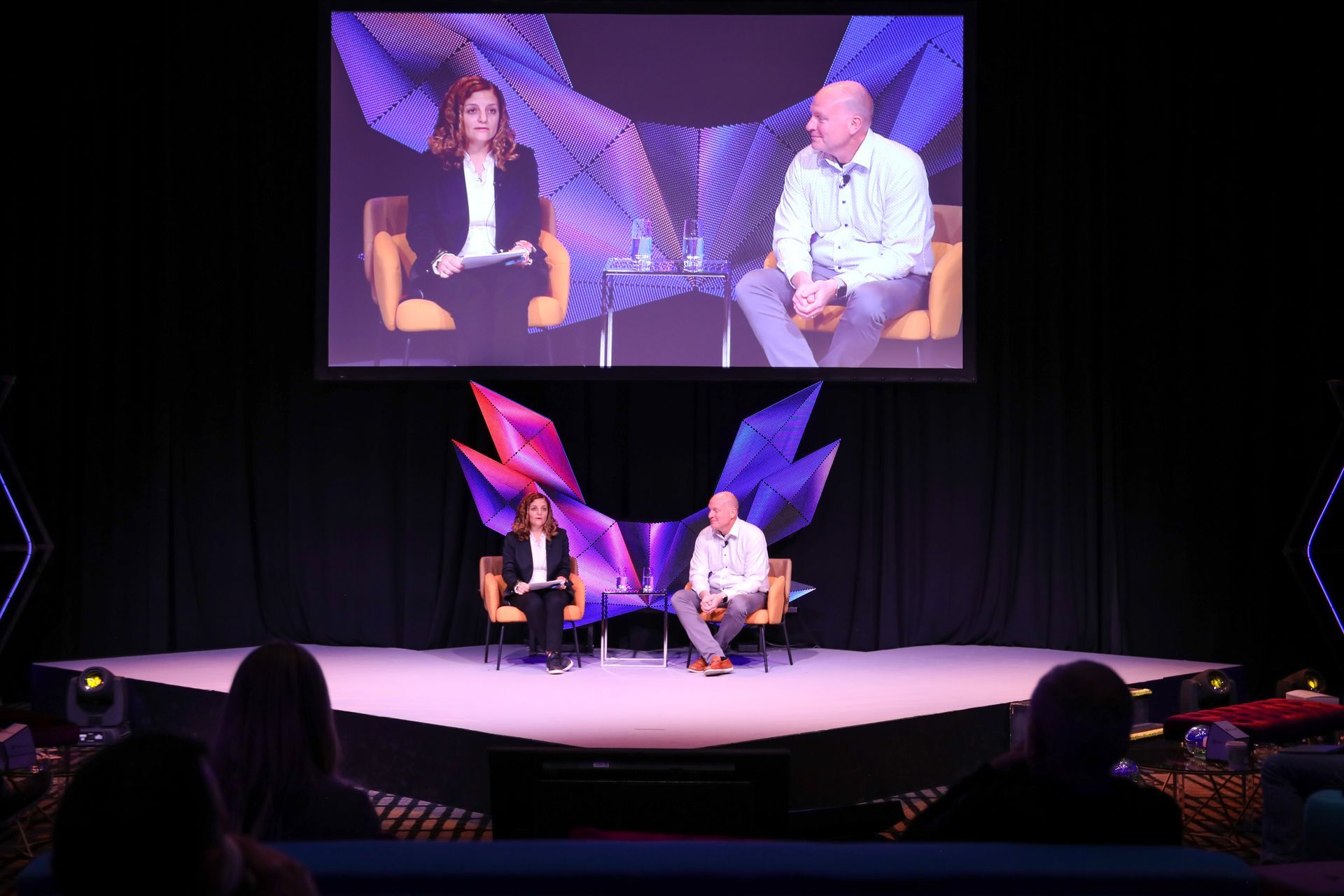 Two people on stage, one interviewing the other, backdrop screen with matching imagery. Audience in seats.