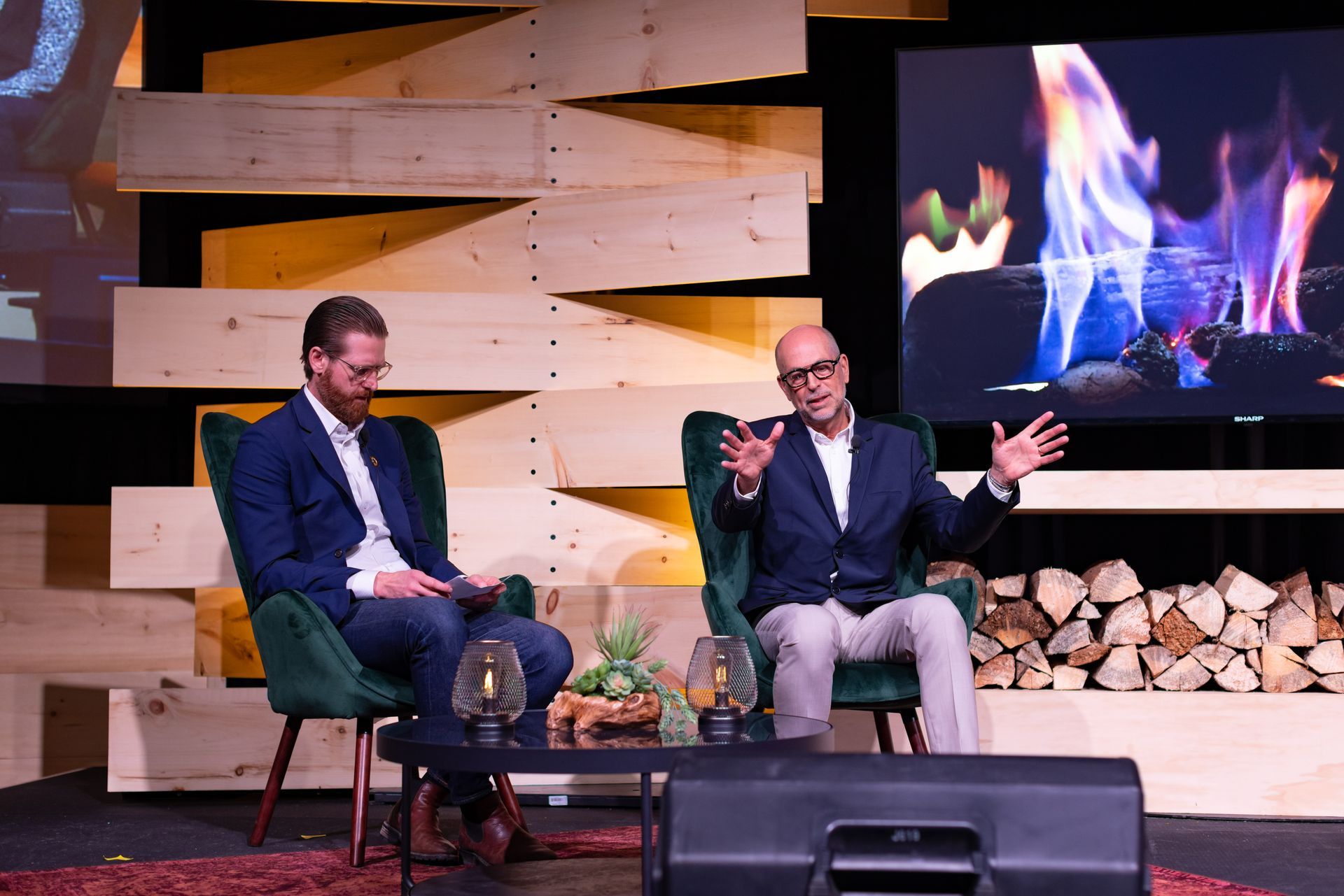 Two men seated on a stage, discussing. One gestures, the other listens. Backdrop features wood and a fireplace projection.