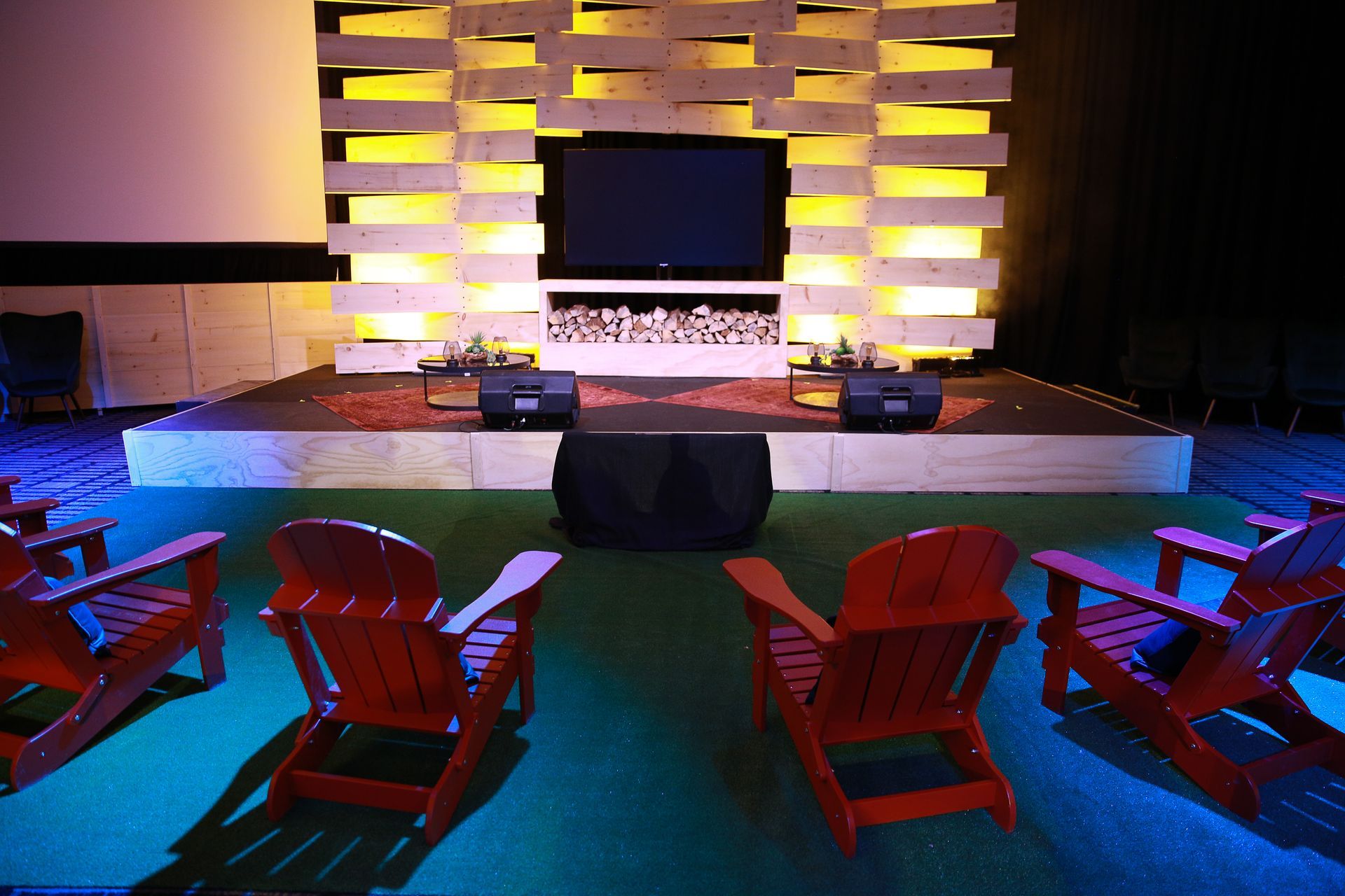 Red Adirondack chairs face a stage with a TV, wood backdrop, and speaker system.