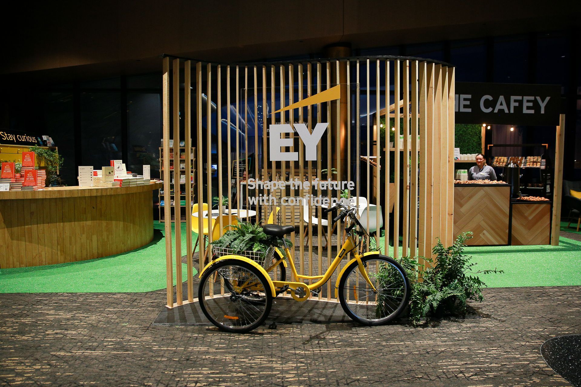 EY-branded display with a yellow bicycle, wooden slat backdrop, and The Cafey sign.