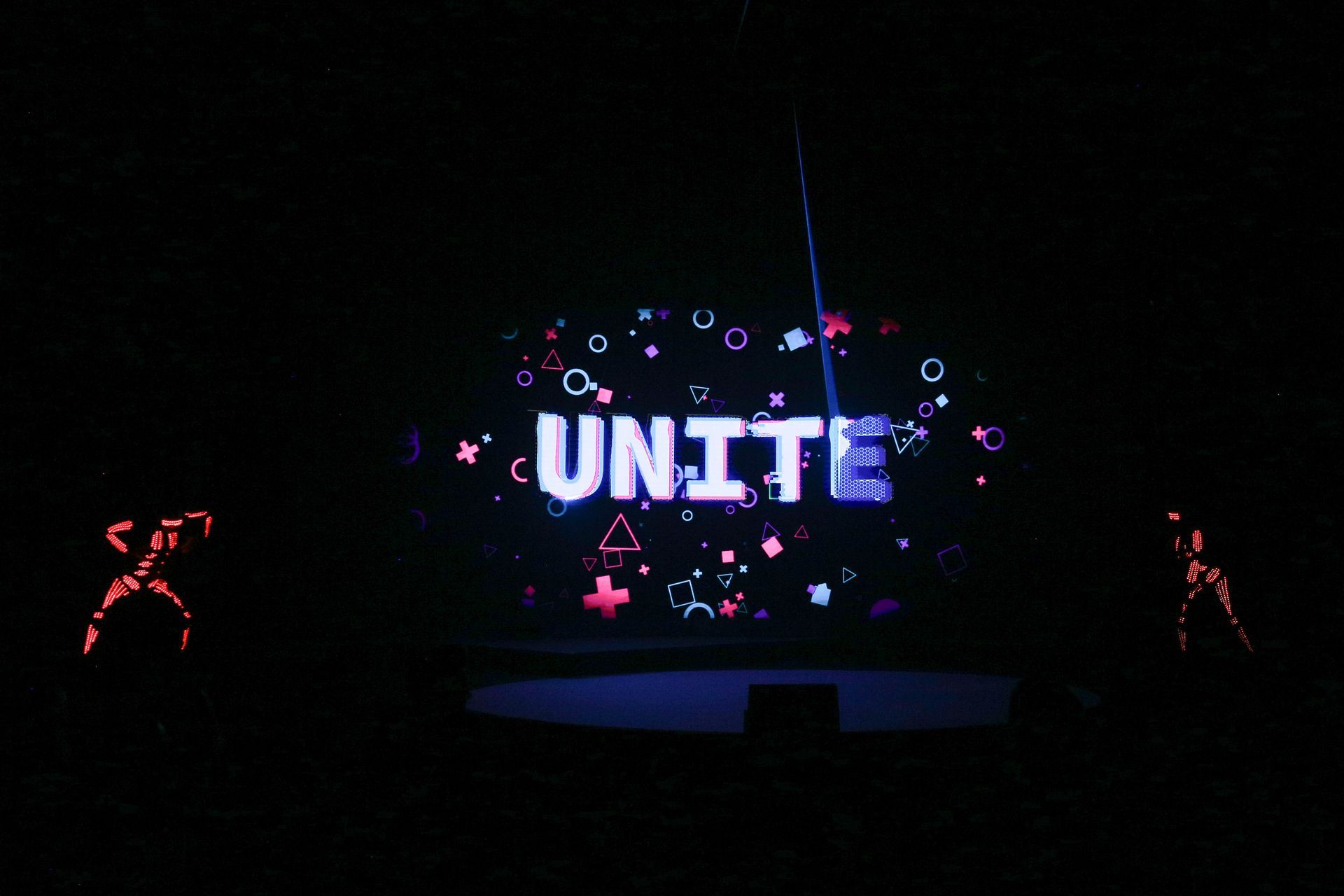“UNITE” lit up in neon on a stage with glowing geometric shapes, two outlined figures, and a dark background.