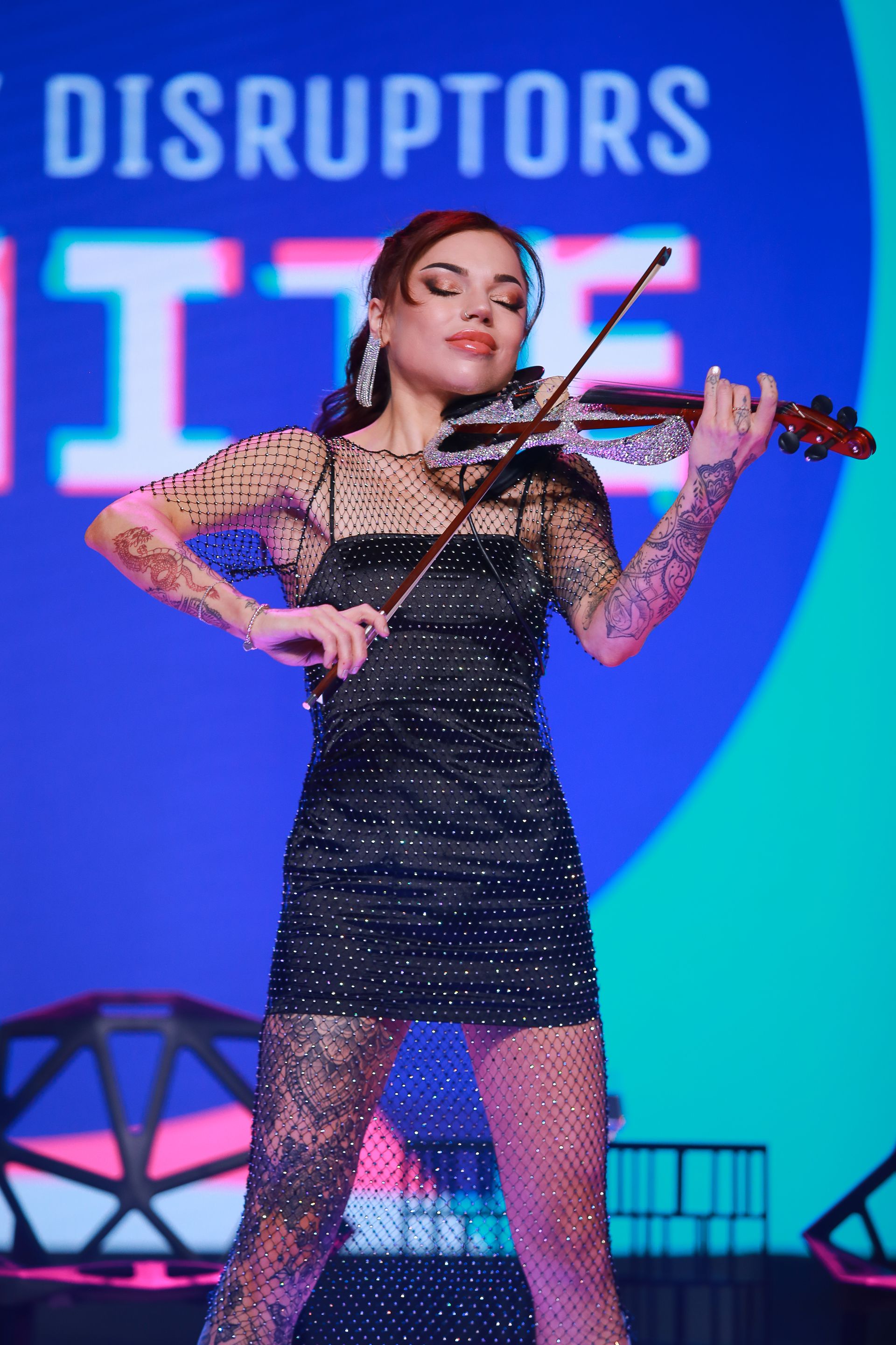 Woman playing a violin on stage, wearing a black dress with mesh details, against a blue backdrop.