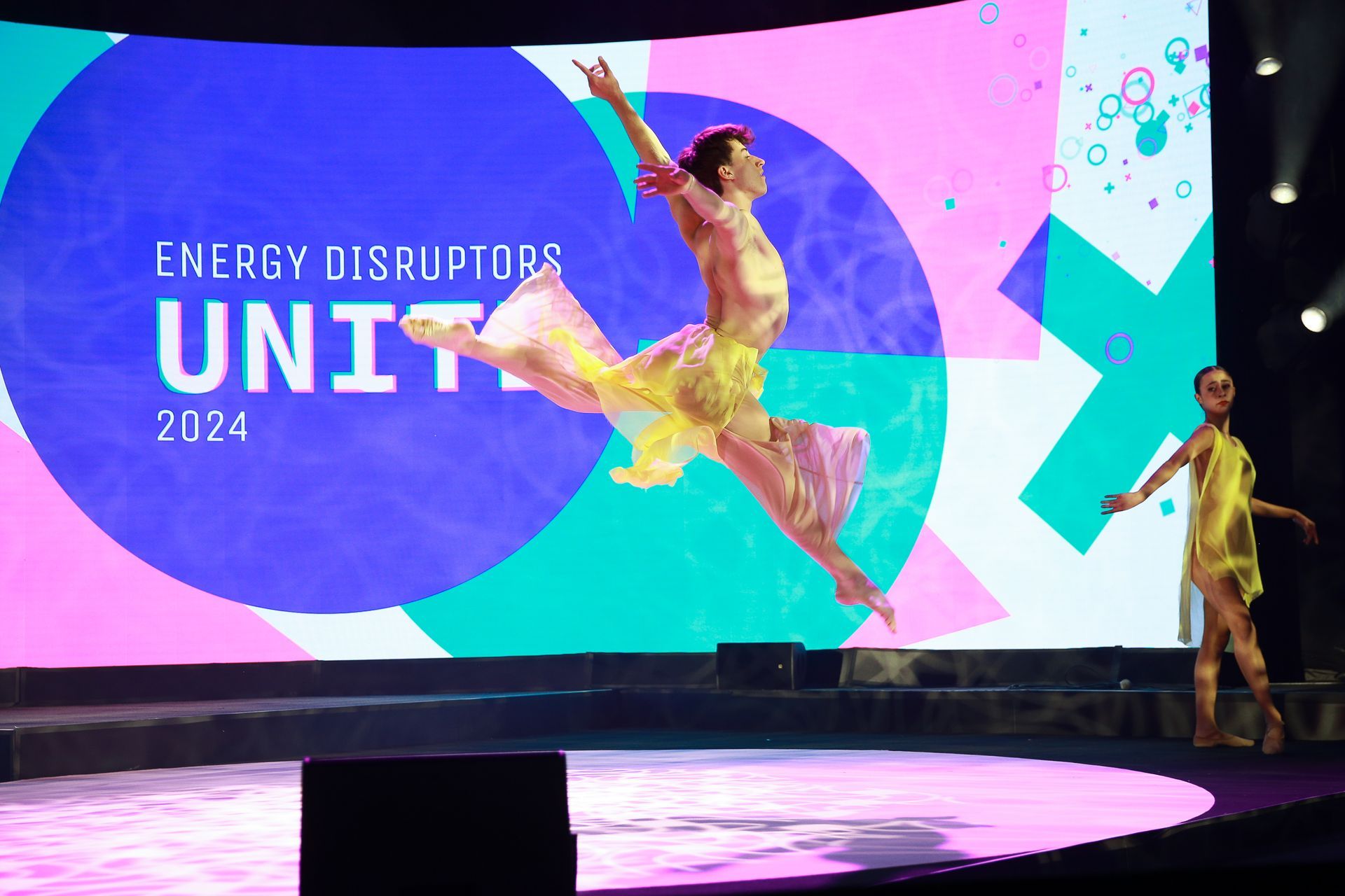 Two dancers on stage, one leaping in yellow dress, event backdrop 